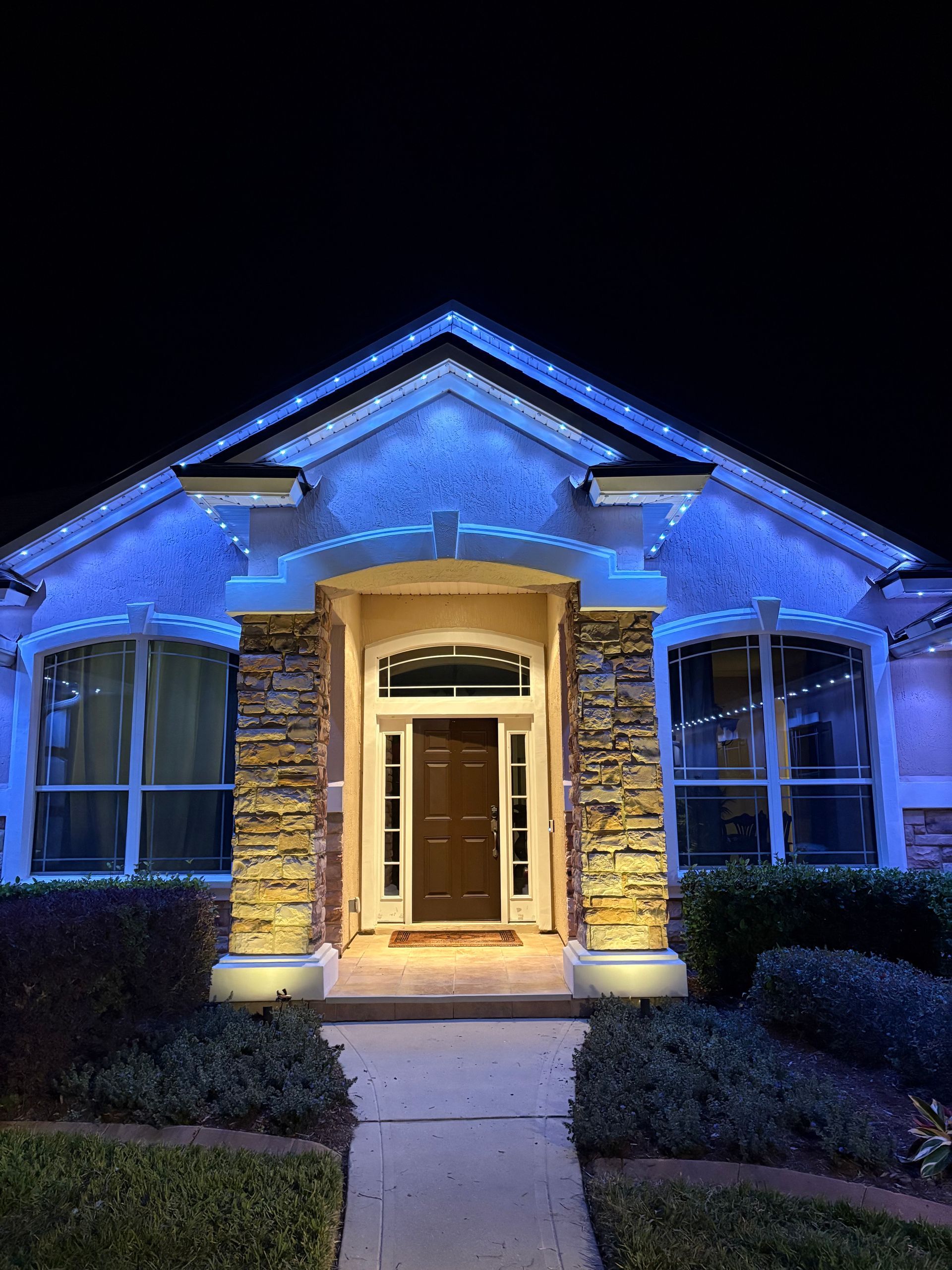 House exterior at night, lit with blue and gold lights. Front door, windows, and landscaping.