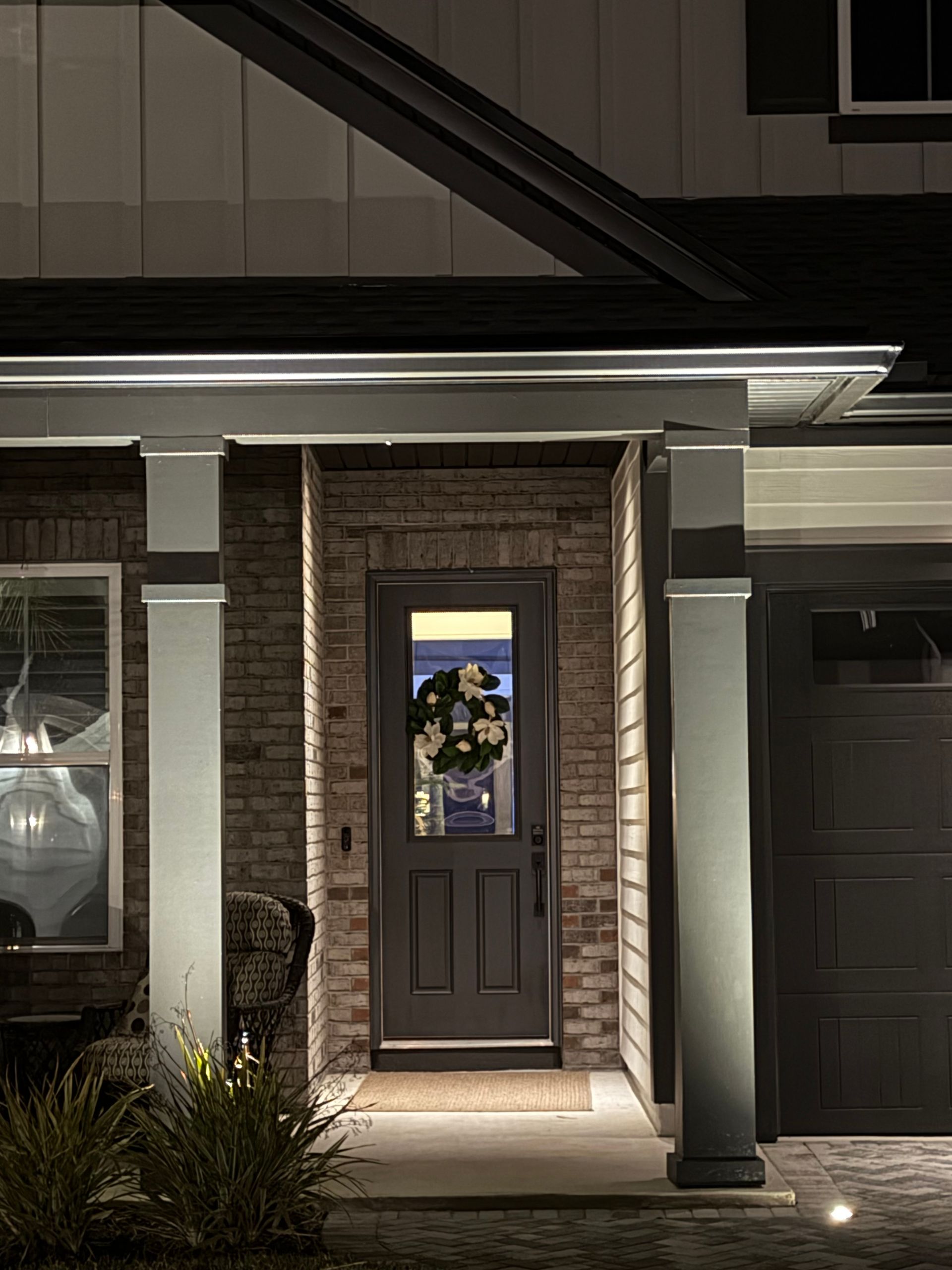 Nighttime exterior view of a home's entryway with illuminated trim. Dark door, brick, and pillars.