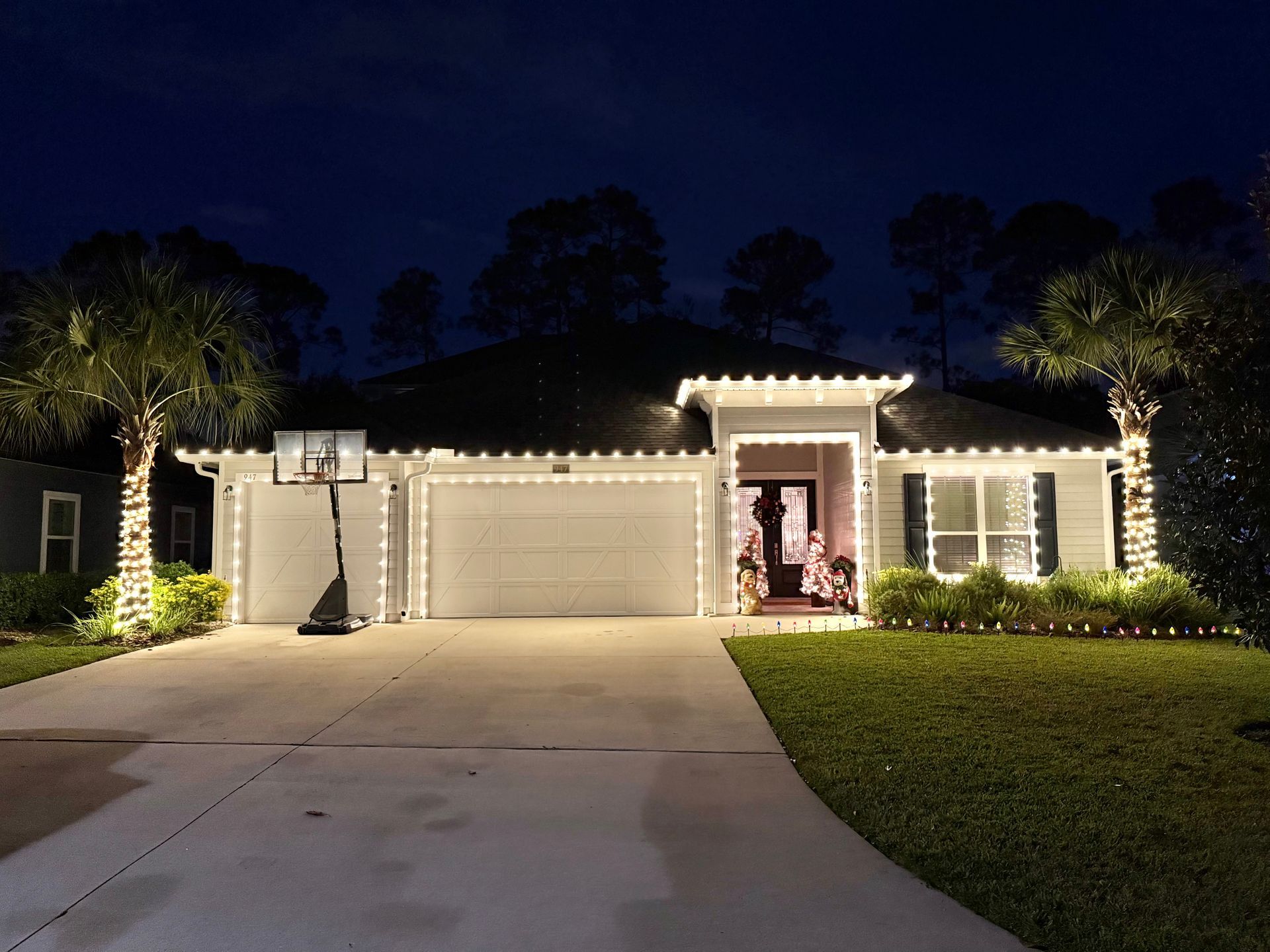 House decorated with white Christmas lights at night; driveway, lawn, and palm trees.