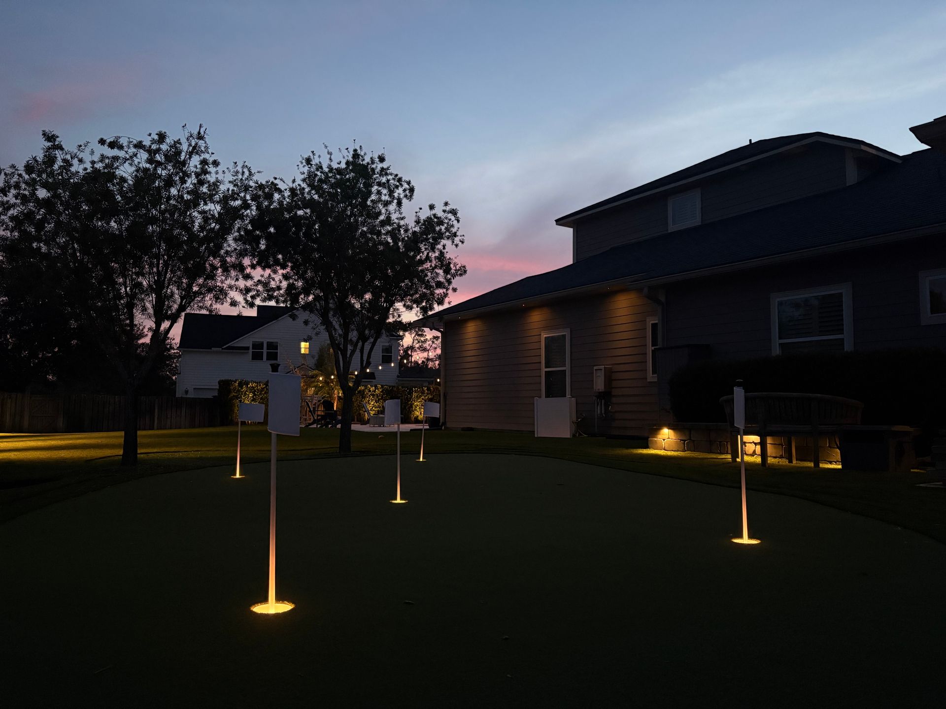 Backyard putting green illuminated at dusk with a house and trees in the background.