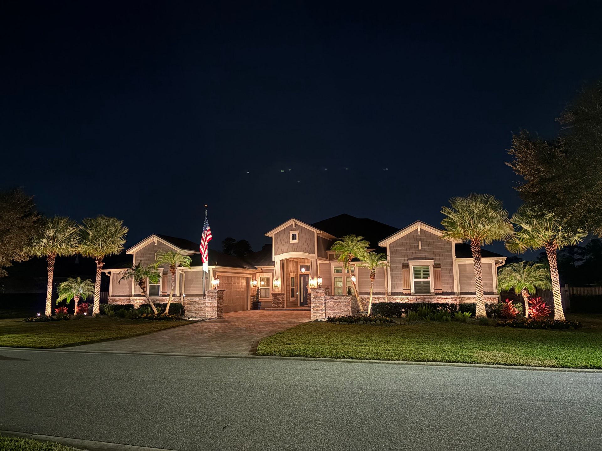 Night view of a house with lit-up features, palm trees, and an American flag.