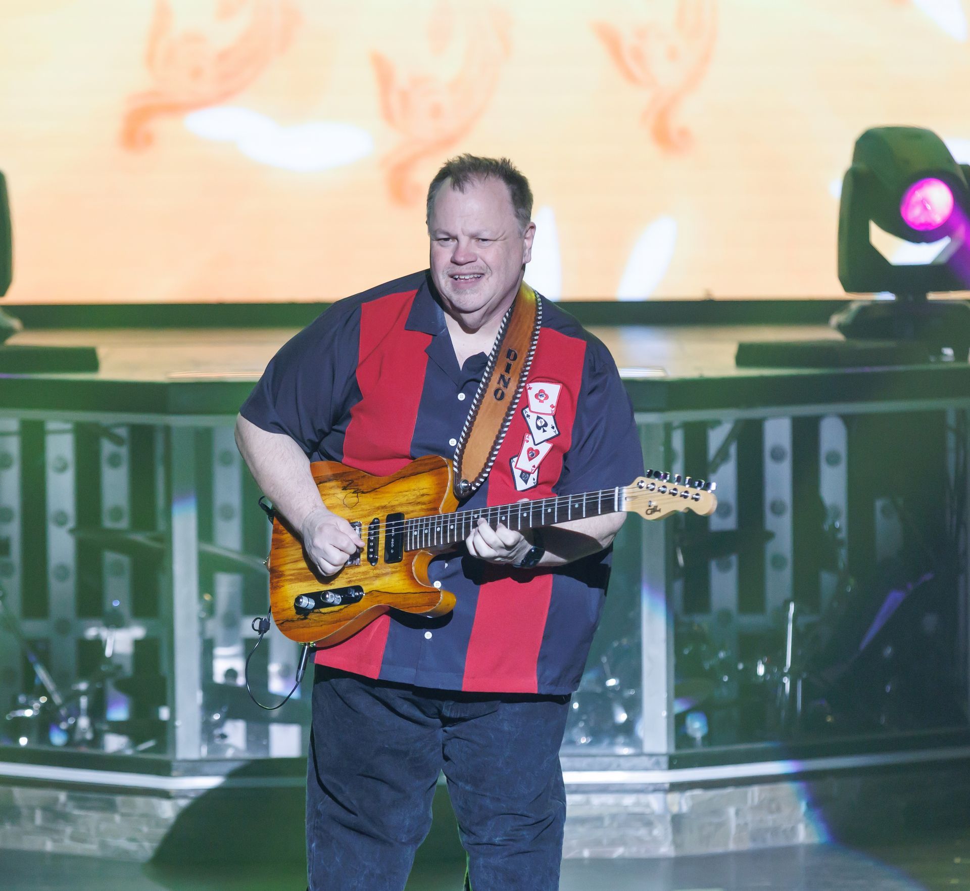A smiling person in a black shirt and red tie plays a bright orange electric guitar on stage next to a microphone.