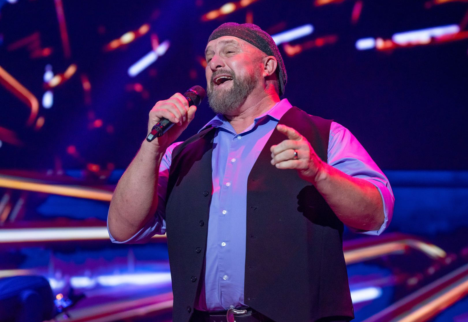 Man in a dark vest and beanie singing into a microphone on a stage, backed by a band with a guitar player.