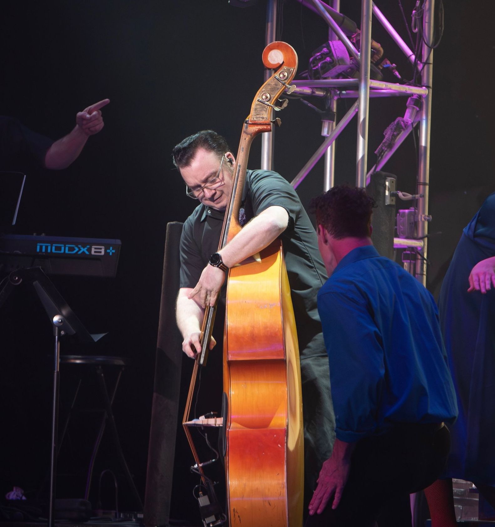 Man plays upright bass onstage; another man kneels nearby. Dim, stage setting; a keyboard is visible.