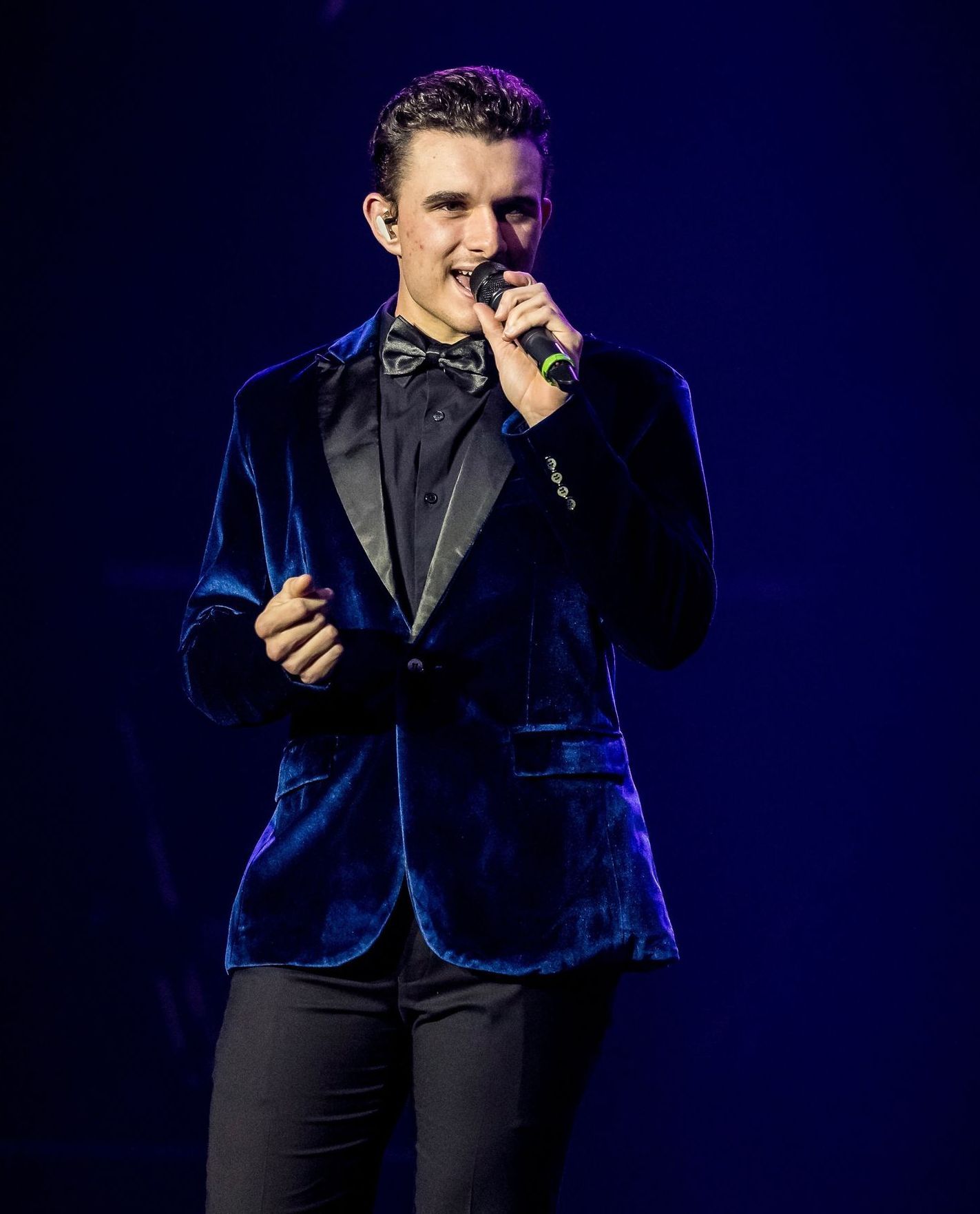Man in a dark blue velvet jacket, black shirt, and bowtie singing into a microphone on stage.