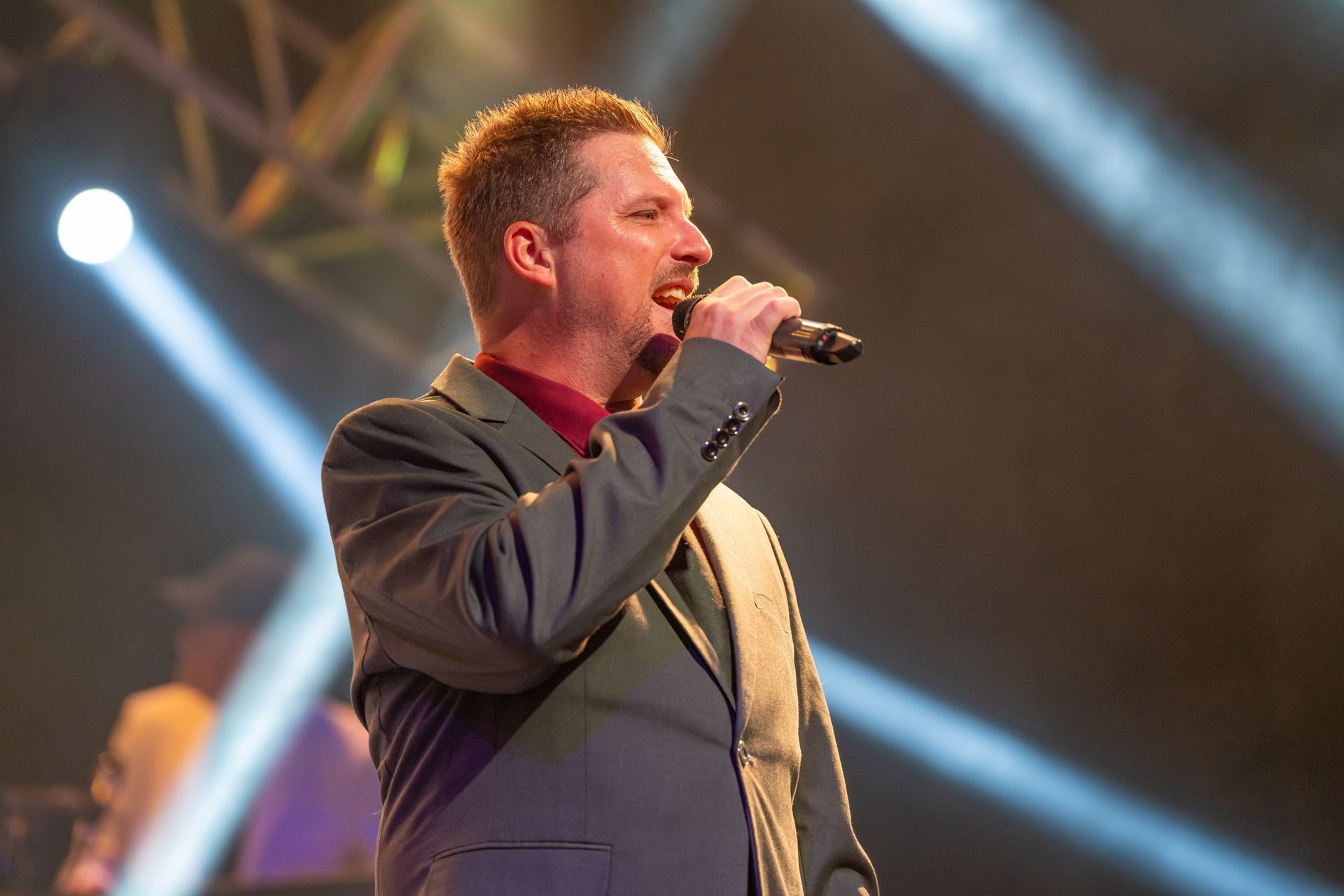Man singing into a microphone on stage, wearing a gray suit jacket and burgundy shirt.