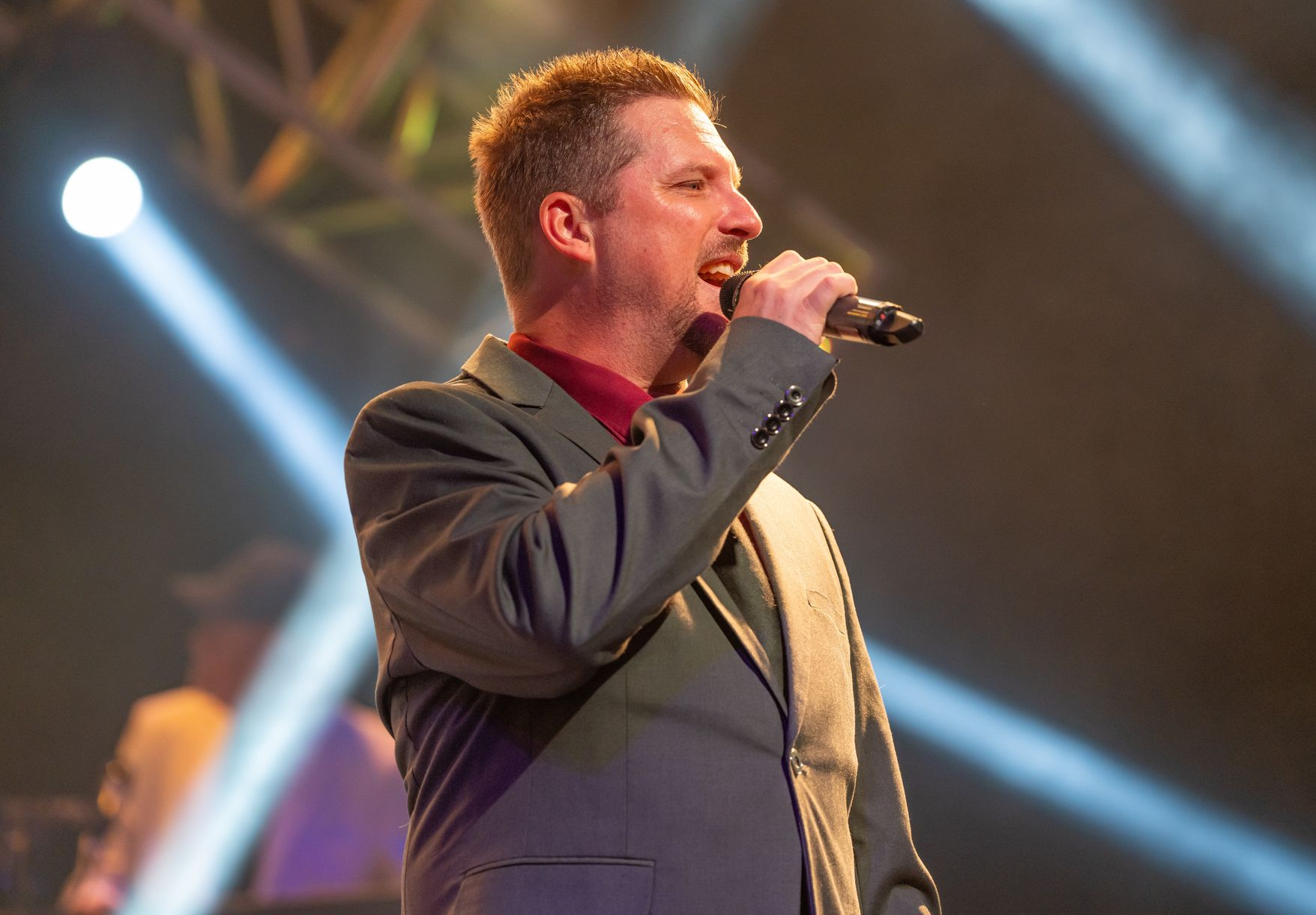 A man in a red blazer and jeans sings into a microphone on stage with a blue background.