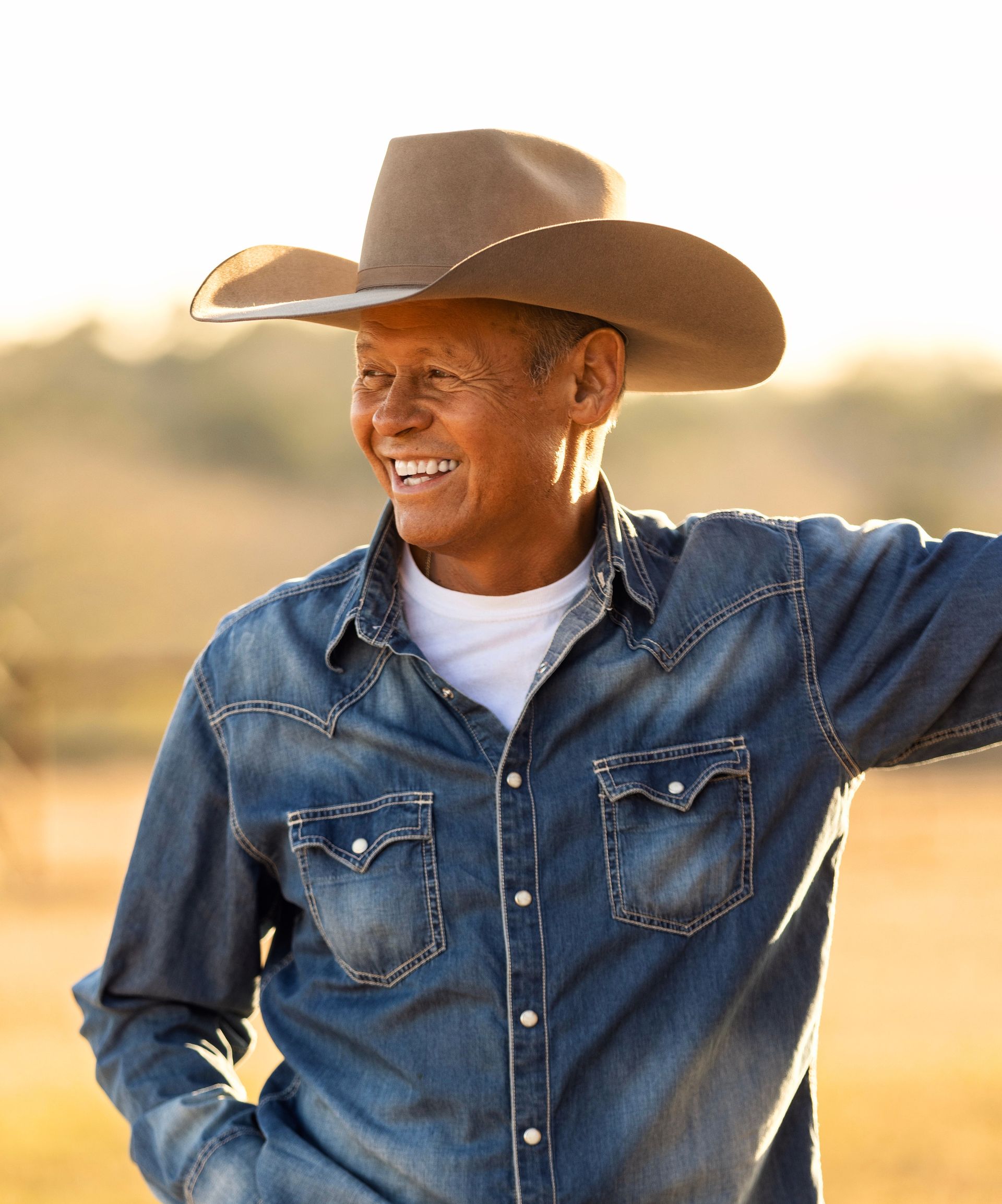Smiling Black man in cowboy hat and denim shirt, outdoors.