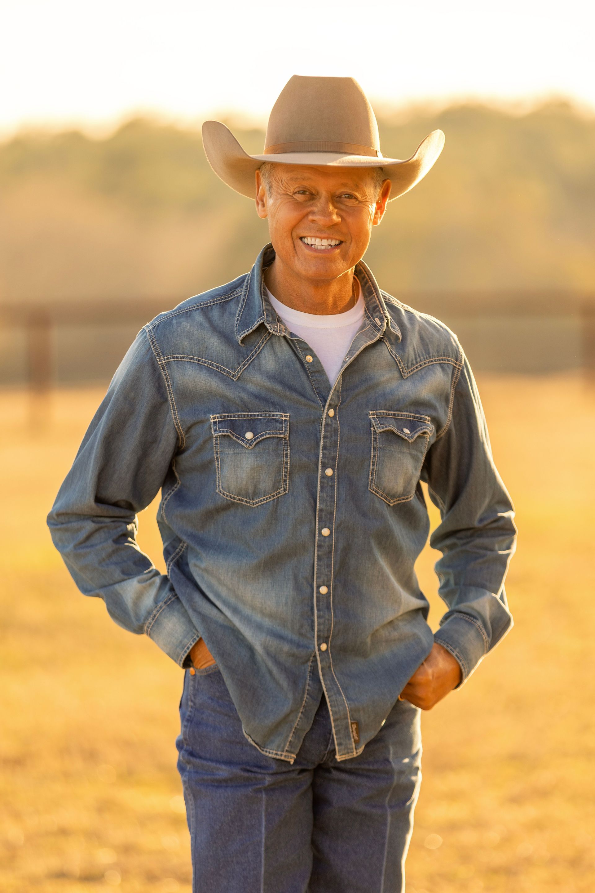 Man wearing cowboy hat and denim shirt smiles outdoors, hands in pockets.