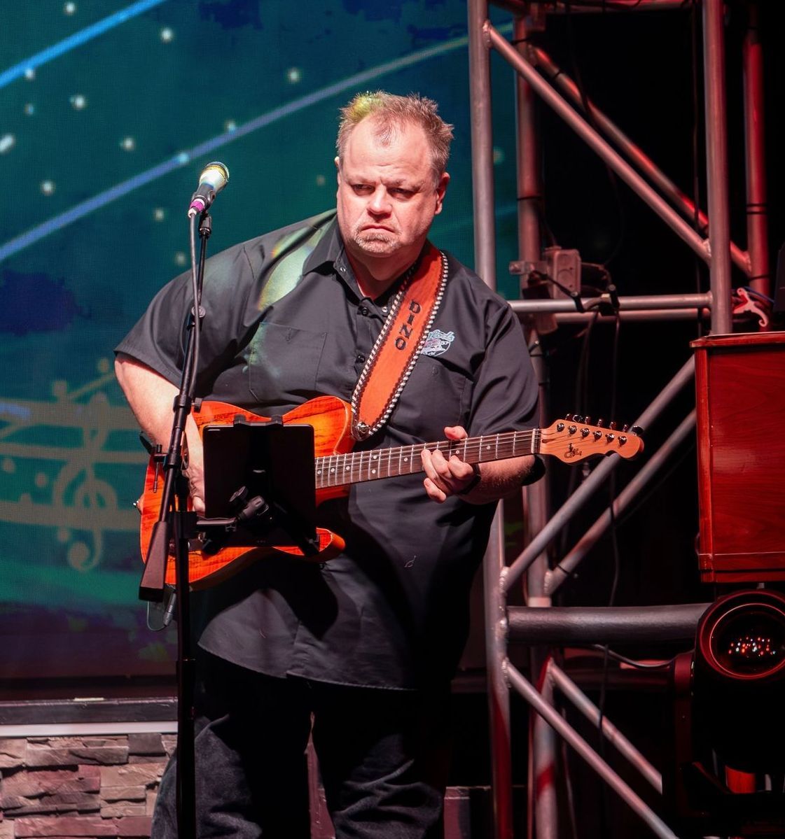 Man playing an orange electric guitar on stage, wearing black shirt.
