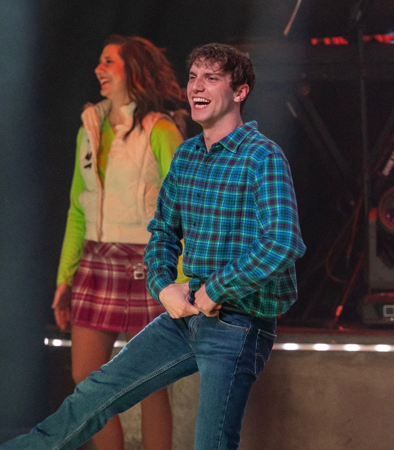 Man in blue plaid shirt and jeans laughs and gestures onstage, woman in background smiles in skirt and hoodie.