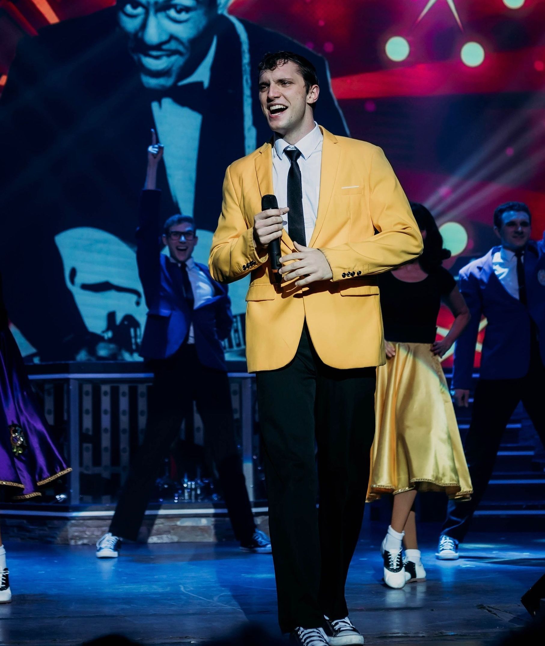 Man in yellow jacket sings on stage, black and white backdrop, dancers in background.