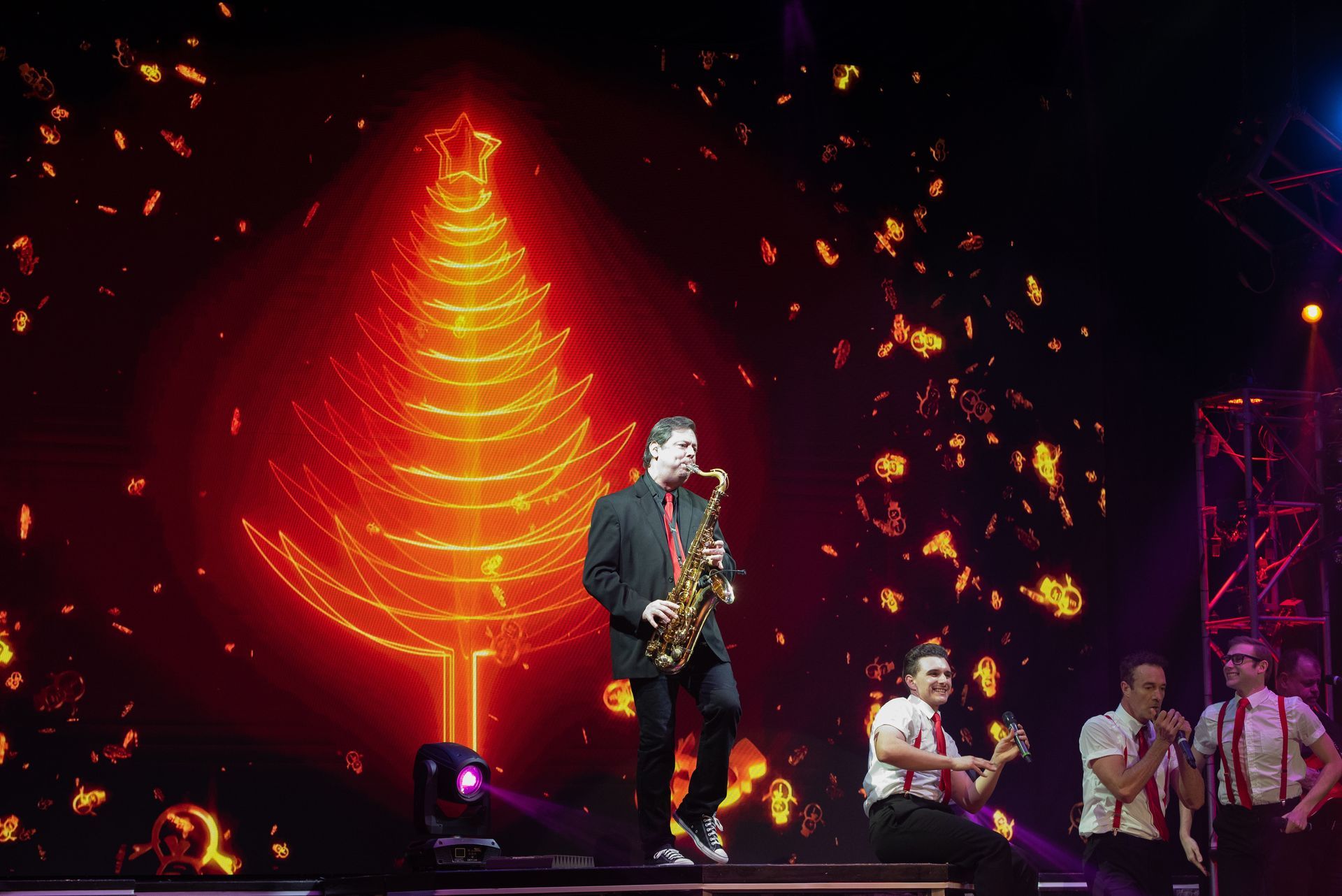 Saxophonist on stage with band, performing in front of a glowing Christmas tree graphic.