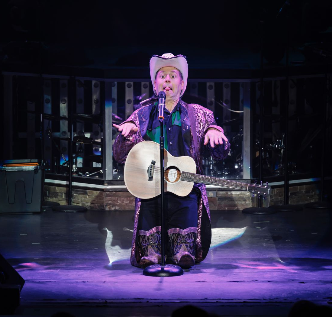 Person on stage in ornate attire, holding a guitar, singing under spotlight.