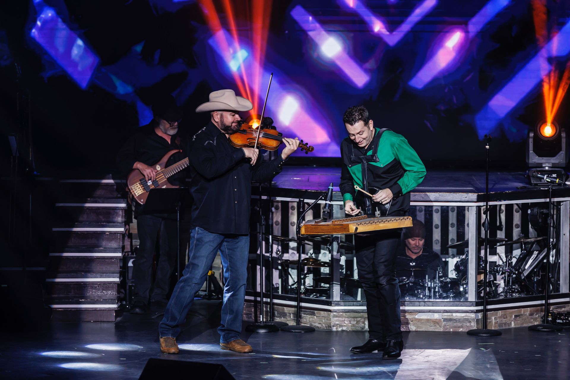 Musicians perform on stage; a violinist and a percussionist play with colorful lights in the background.