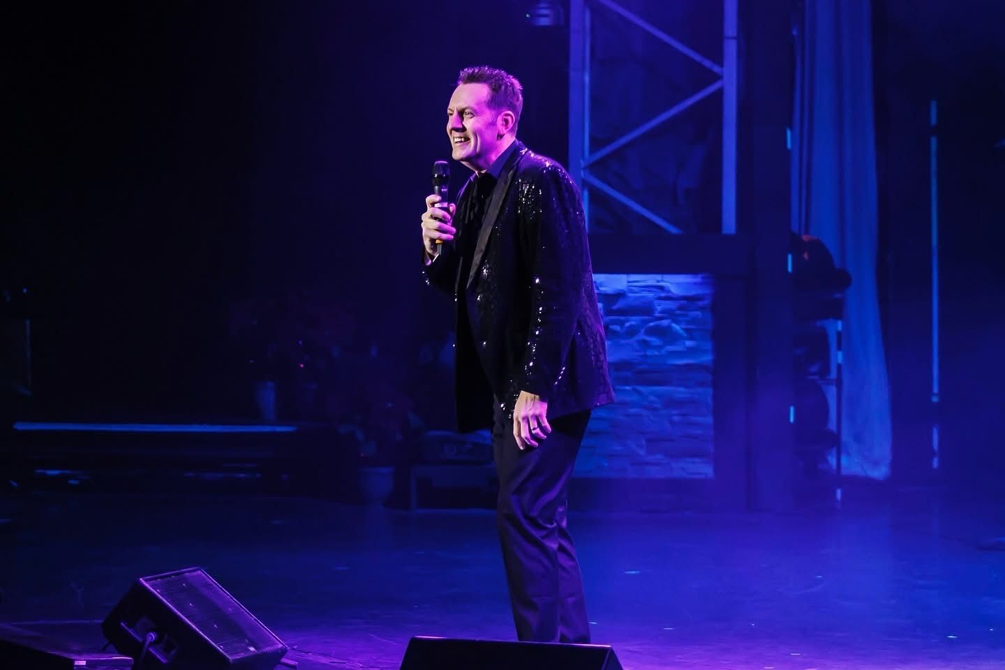 Man in sequined jacket holds microphone on stage, smiling. Blue stage lighting.