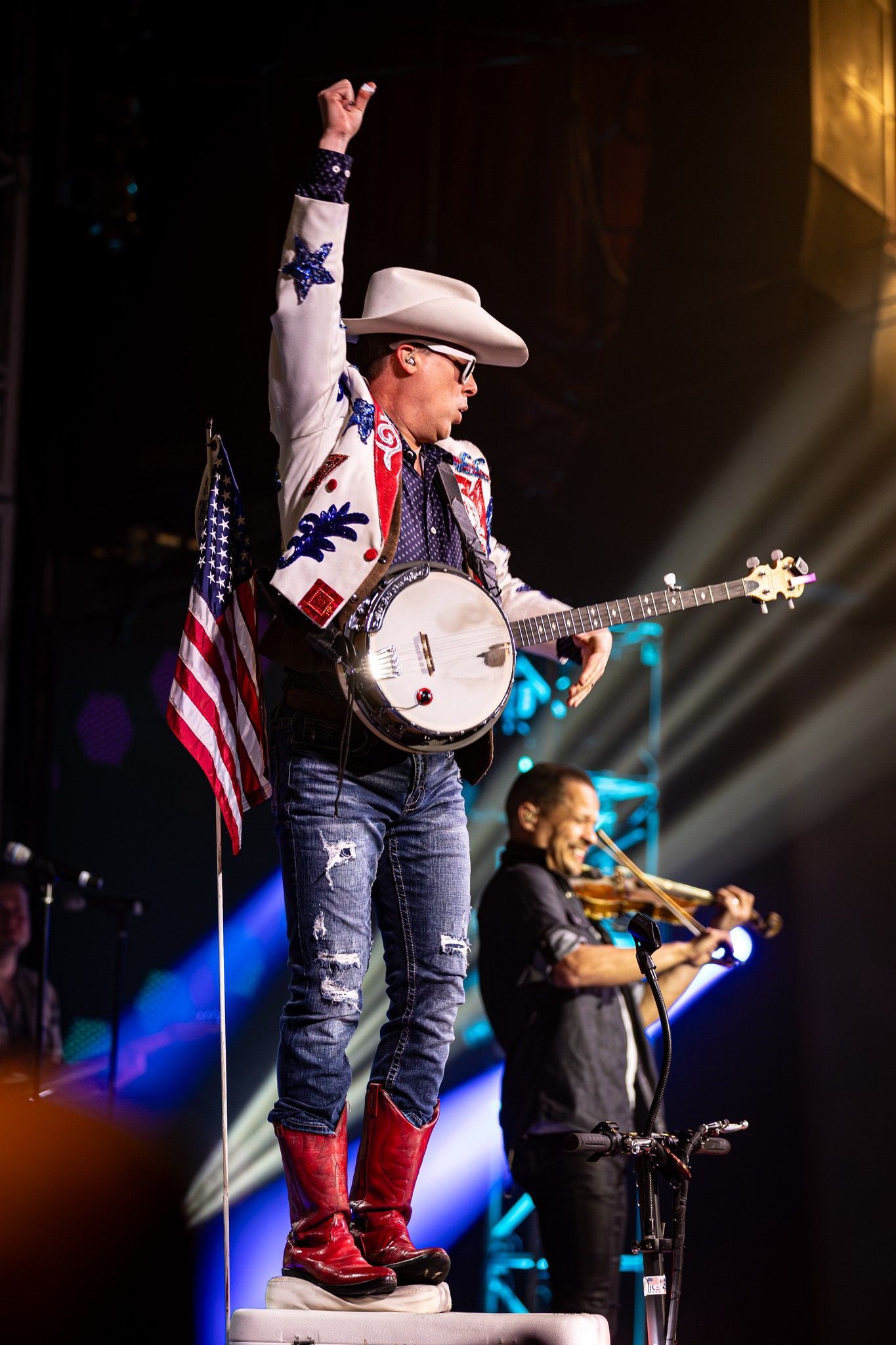 A performer in a white cowboy hat and sequined red, white, and blue jacket plays a banjo while standing on a platform with an american flag behind him and his brother is in the background playing fiddle