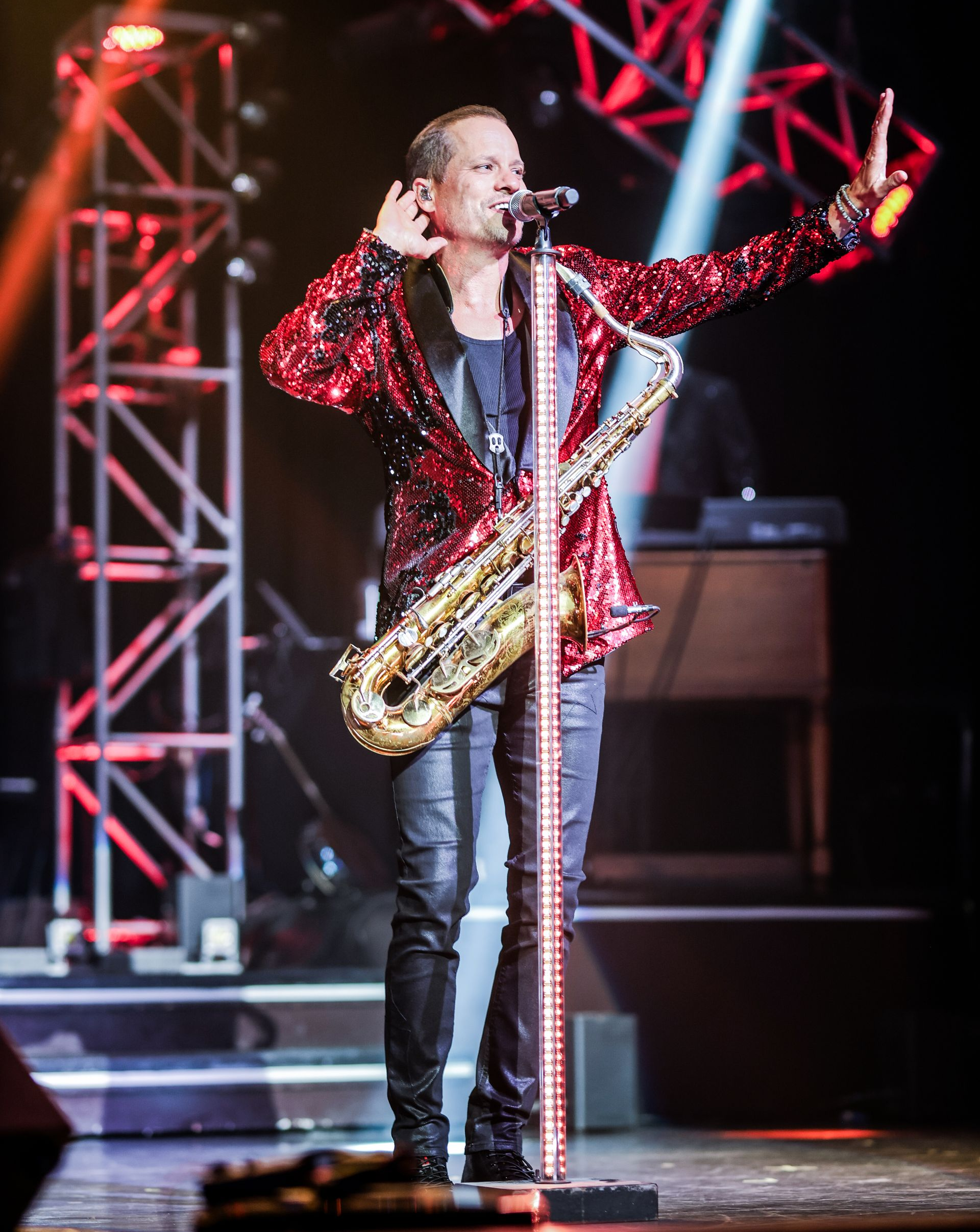 A performer standing at a microphone stand wearing a red sequined jacket playing a saxophone on a stage with his hand out 