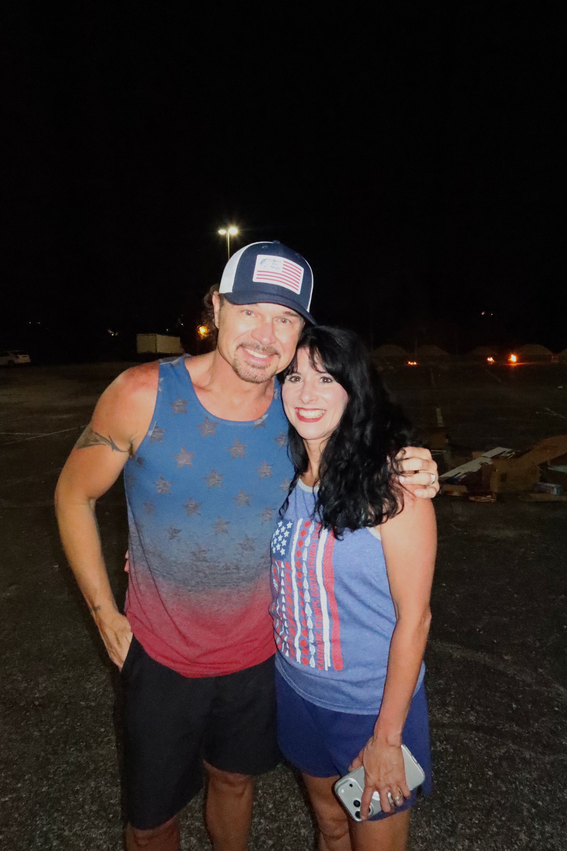man and woman hugging in fourth of july clothes outside in a dark parking lot with a bright flash possibly shooting off fireworks