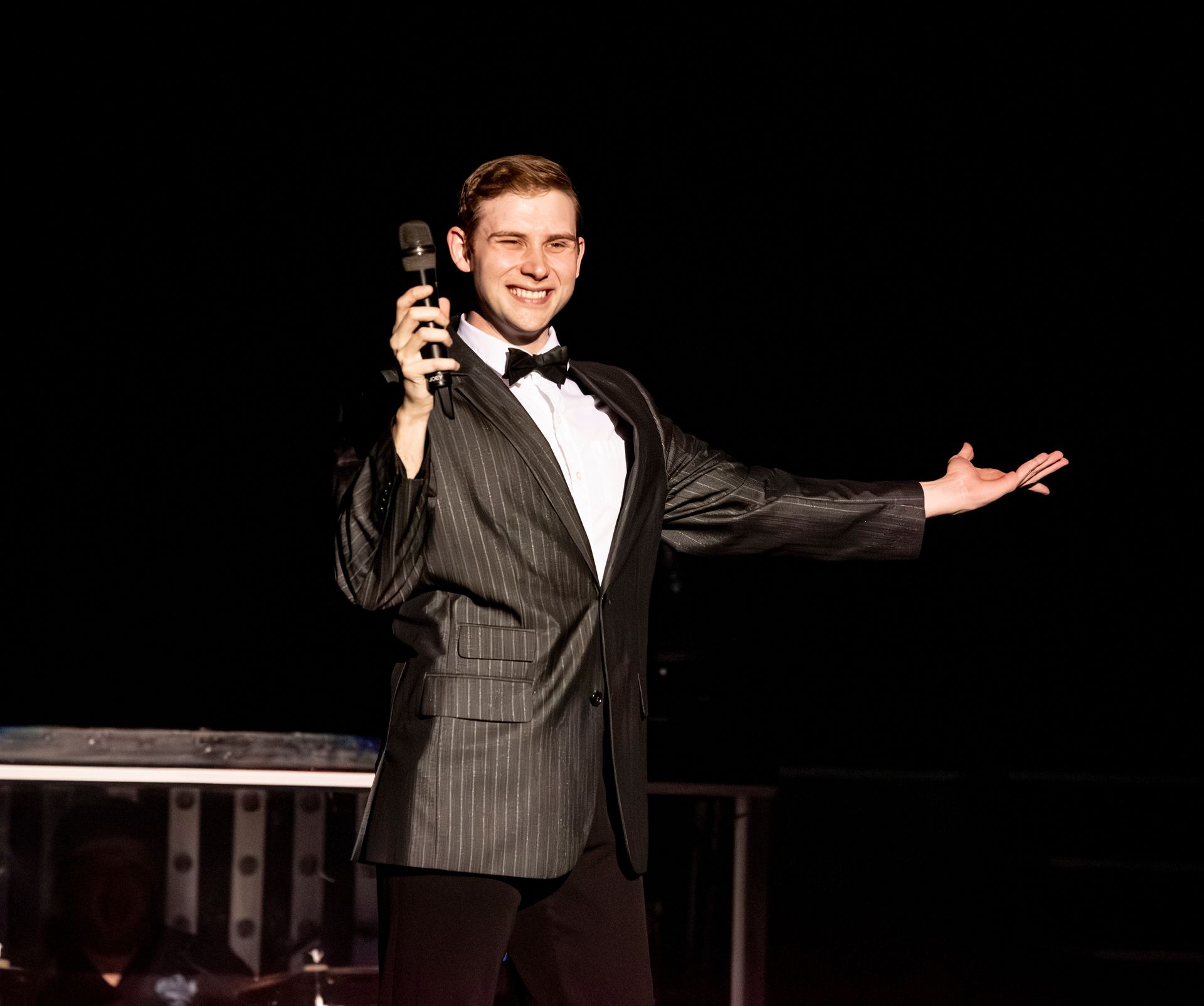 A person in a pinstripe suit and bow tie smiles while holding a microphone on a stage with vibrant lighting.
