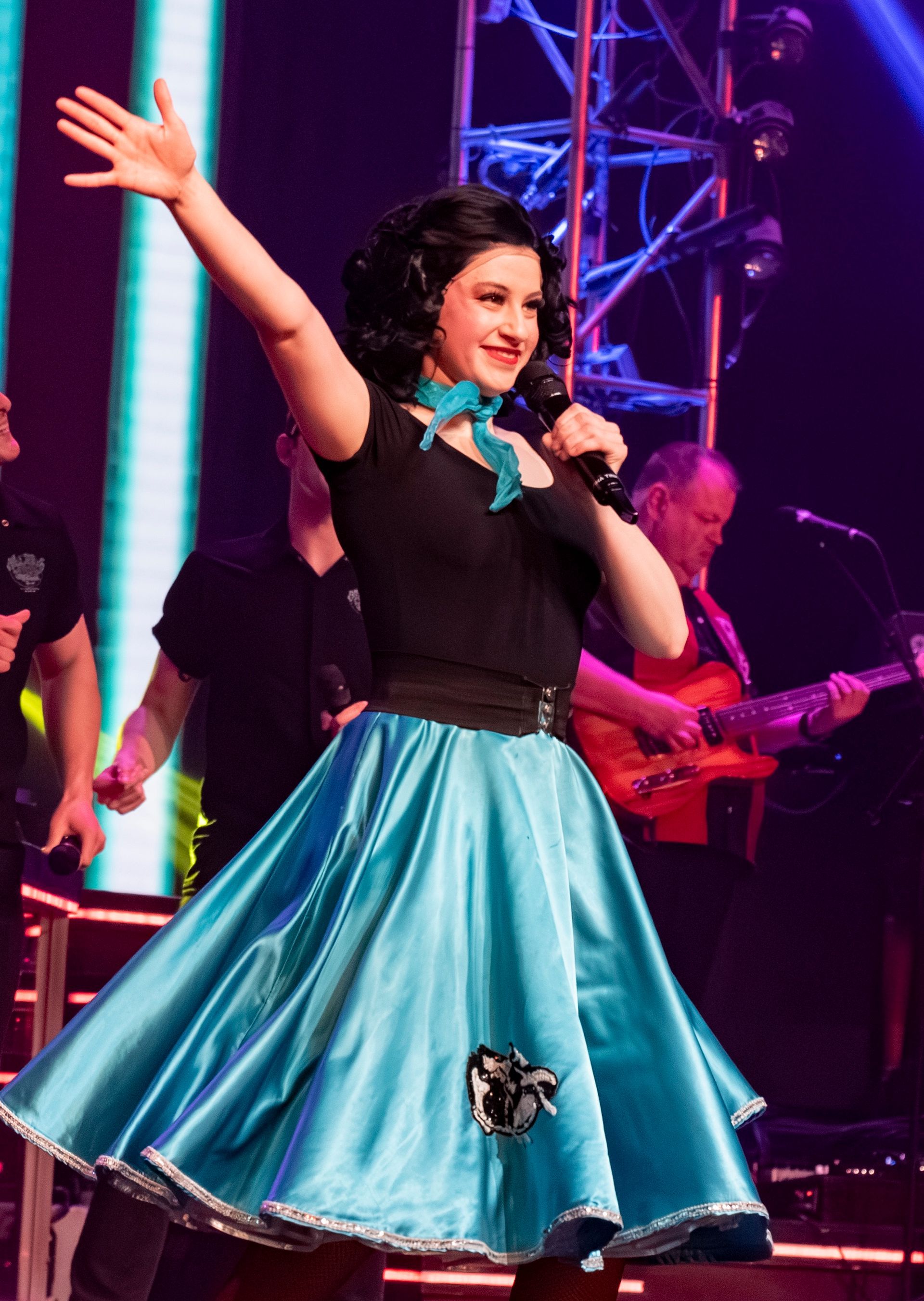 A performer in a black top and blue skirt waves on stage with a microphone 
