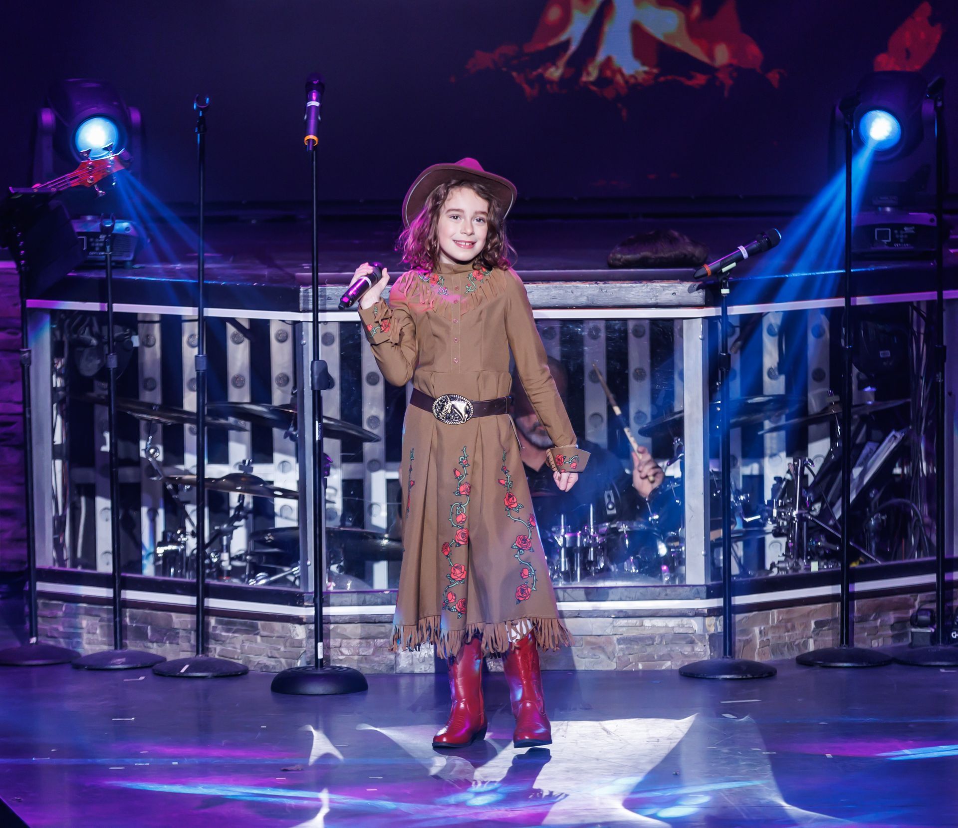 Young singer on stage, wearing a brown outfit with cowboy hat, boots, and holding a microphone.