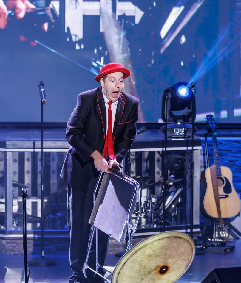 Man in red hat and suit on stage, performing with a cymbal. Band in background.
