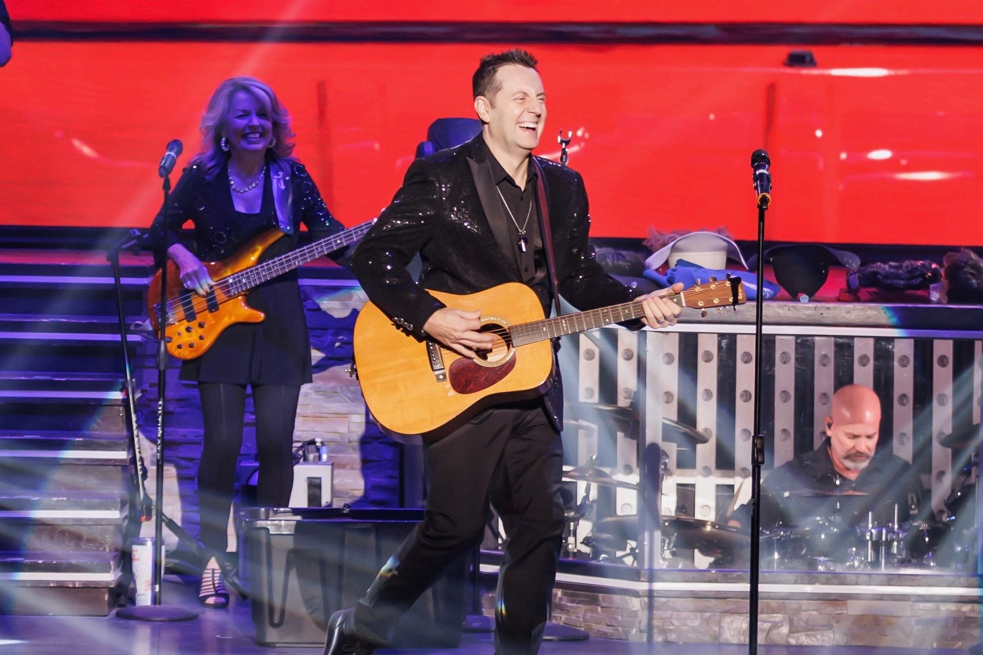 Man in sequined jacket plays guitar on stage with band.