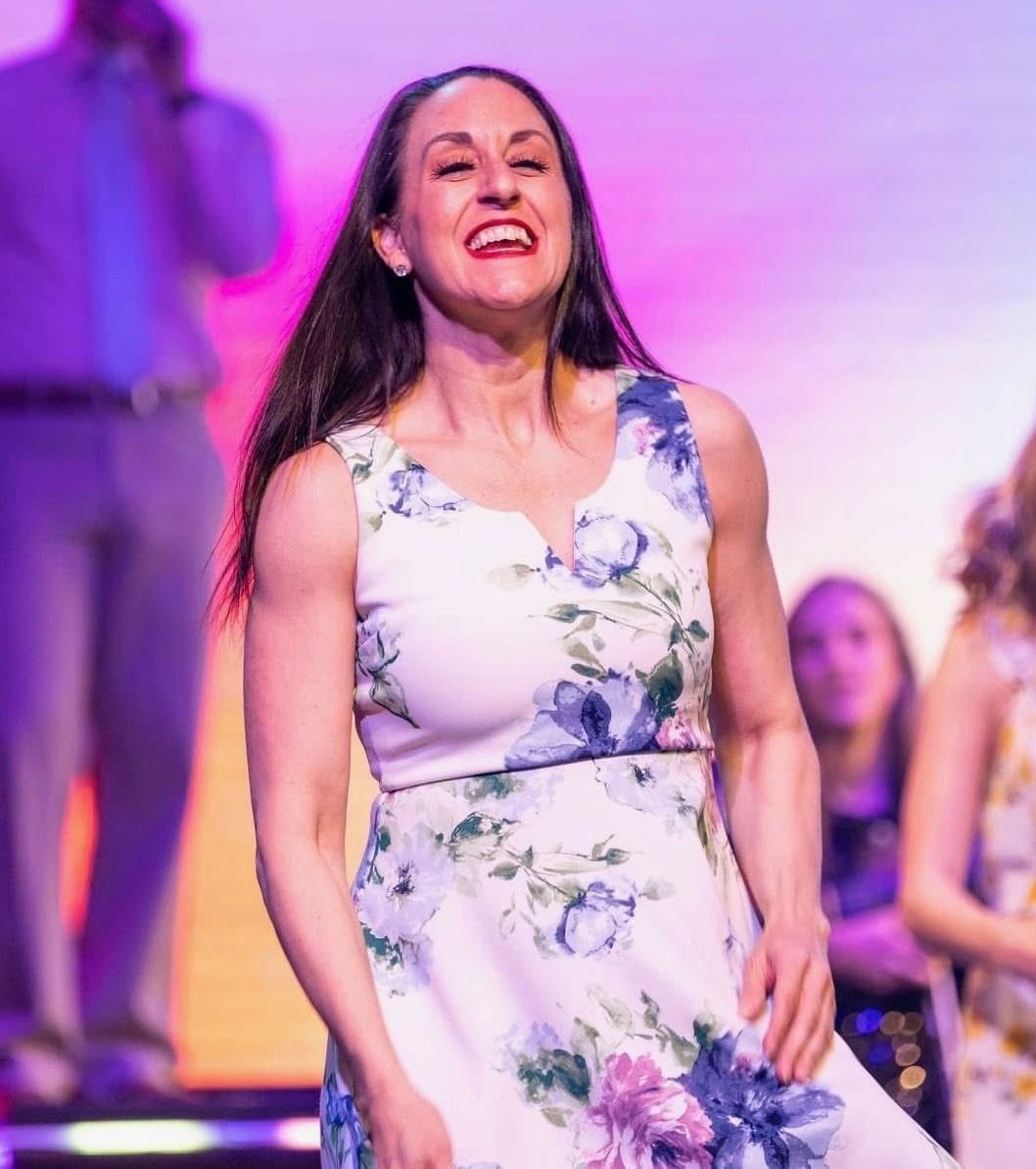 Woman in a floral dress, smiling broadly, on a stage. Backlit with colorful stage lights.