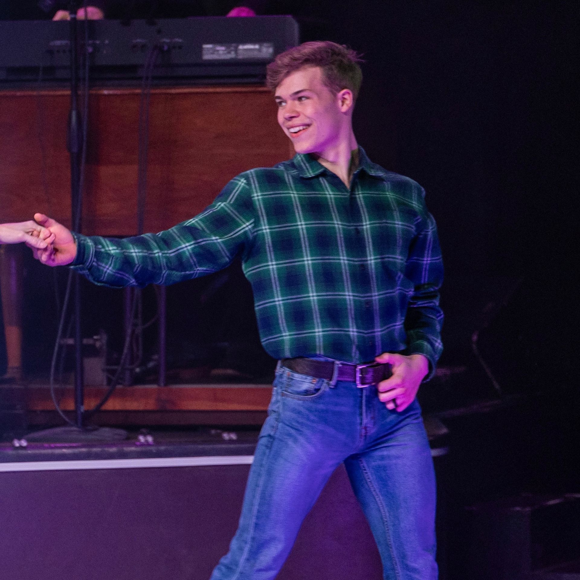 A young man dances on stage, wearing a green plaid shirt and blue jeans. He smiles, extending his arm to the right.