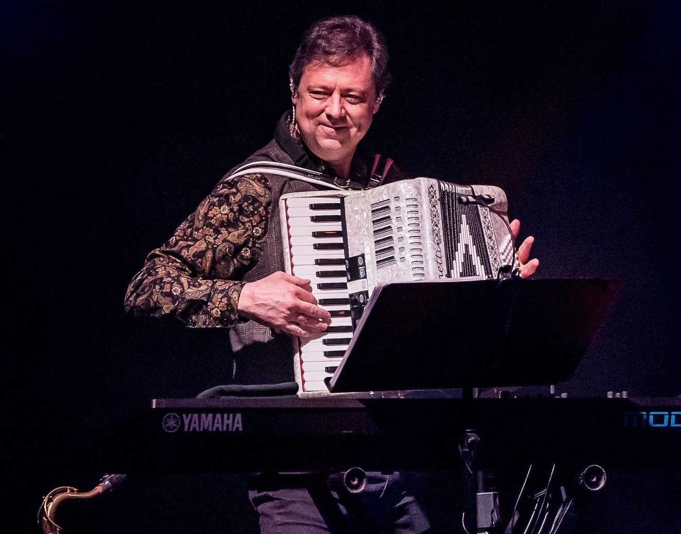 A man plays an accordion and keyboard on stage with a saxophone at his side. He wears a patterned jacket.