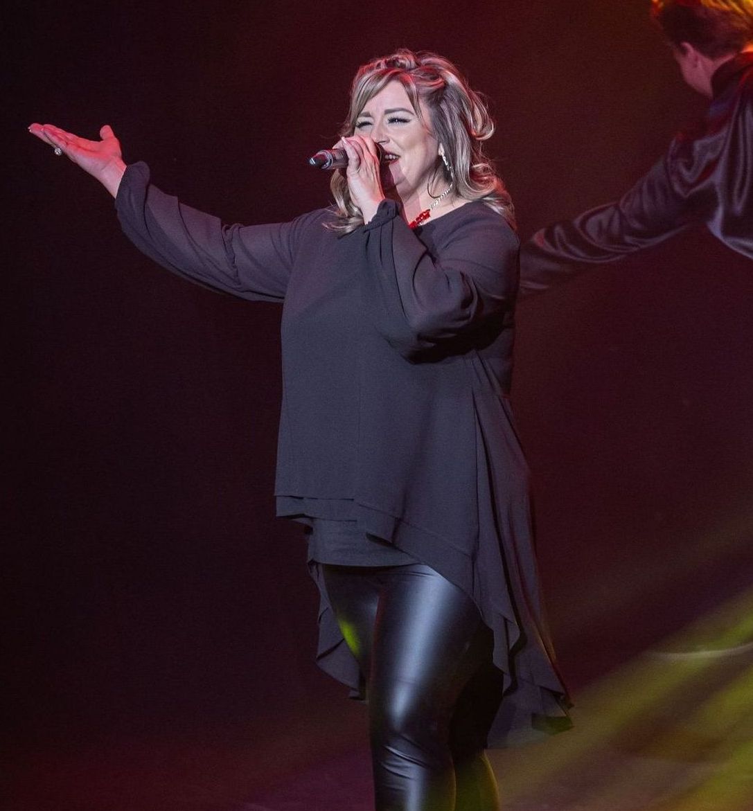 Woman singing on stage, wearing a black top, leather leggings, and red boots, arms outstretched.