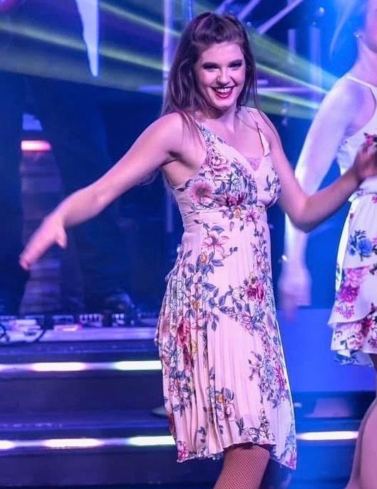 Two women in floral dresses dance on a stage with a band in the background. The woman in front smiles.