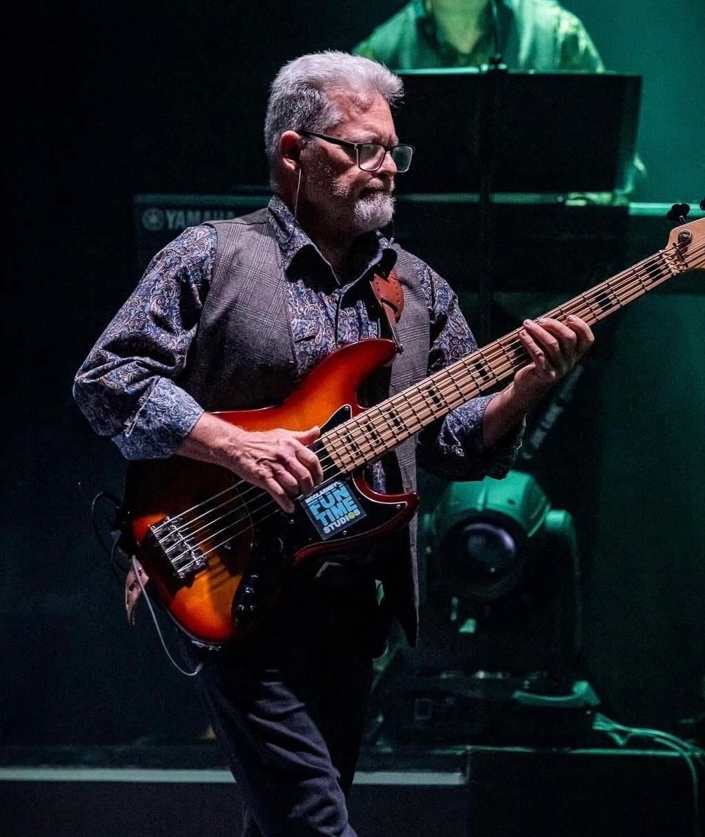 Man with gray hair and glasses playing a bass guitar on stage, wearing a patterned shirt and vest.