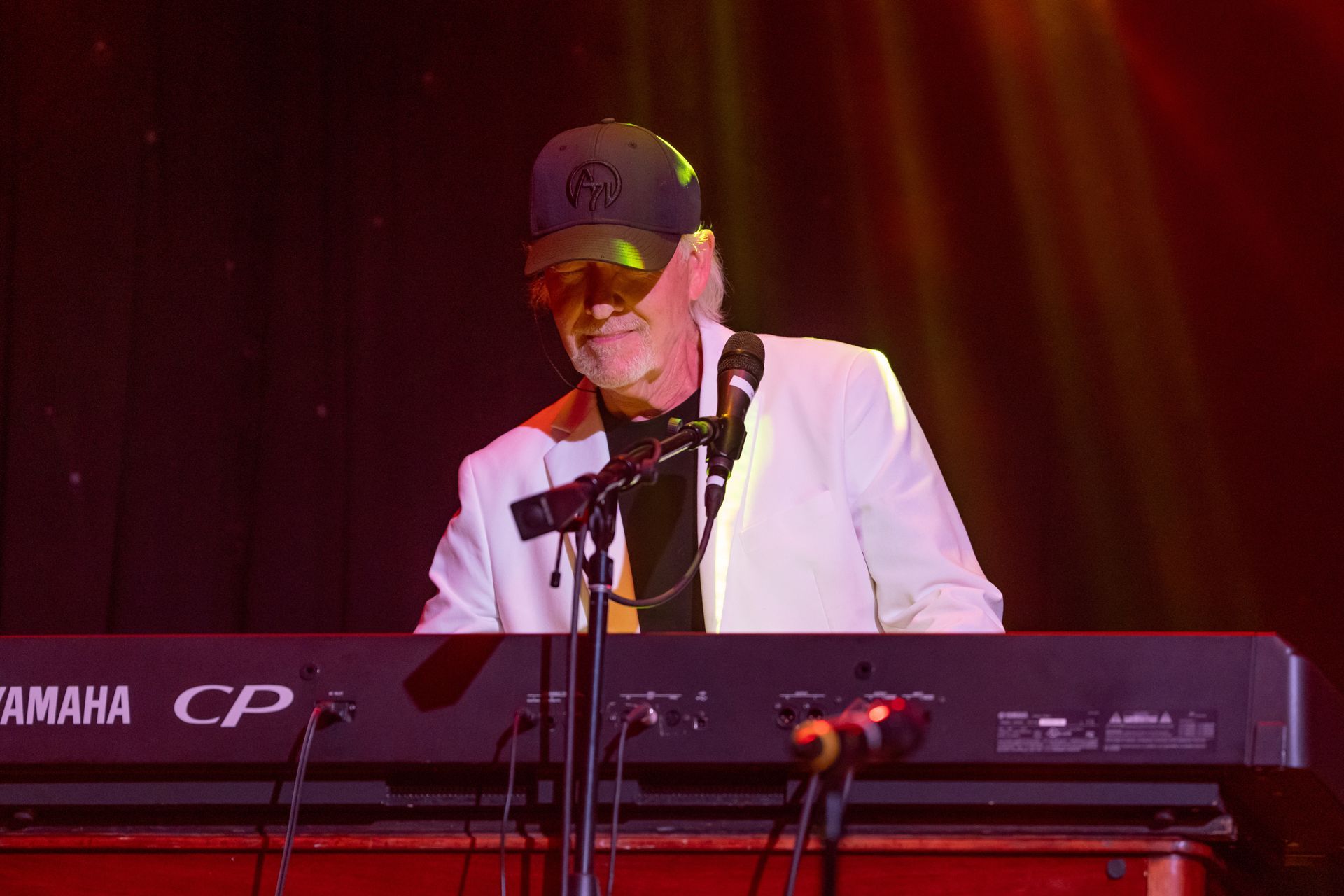 A musician playing a keyboard on stage; he wears a black cap and a white jacket.