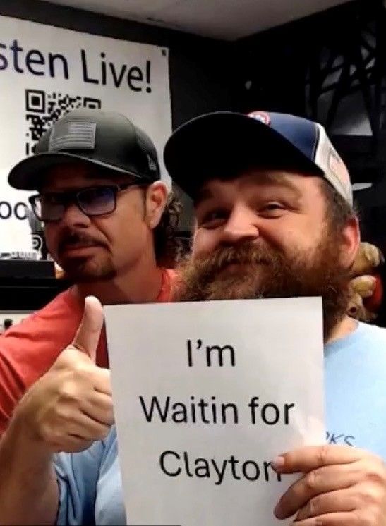 Two smiling men at a protest hold a sign reading “I’m Waitin for Clayton,” one giving a thumbs-up.