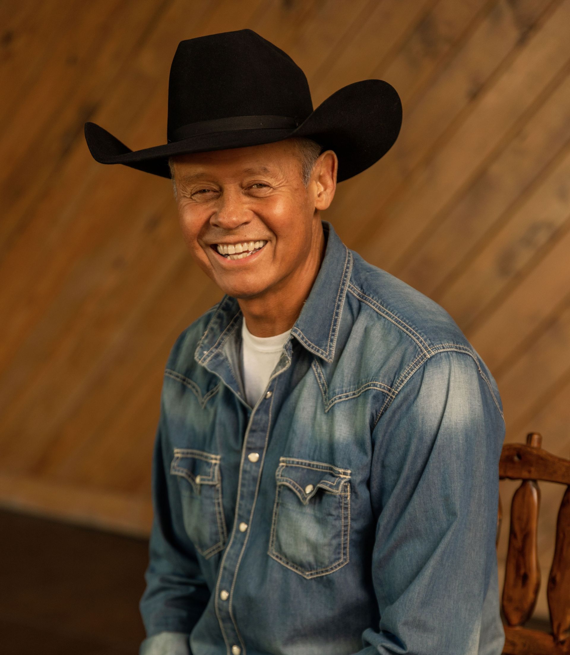 Smiling man in cowboy hat and denim shirt, seated against a wood wall.