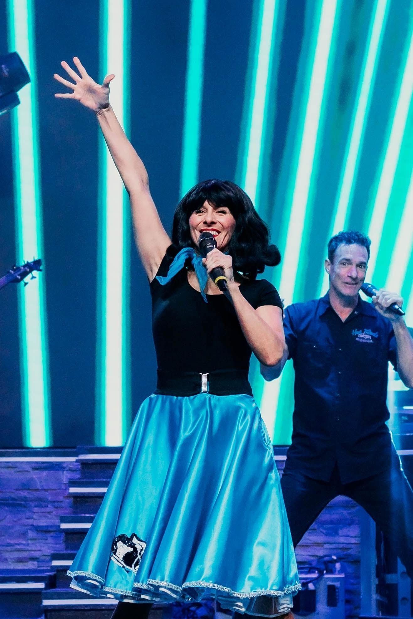 Woman in 1950s attire sings, arm raised, with a man singing beside her on stage. Blue neon backdrop.