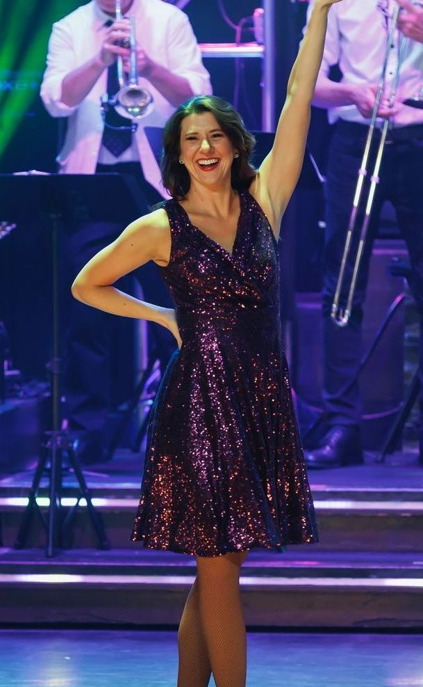 Woman in a sparkly purple dress on stage, arm raised, smiling. Band in the background.