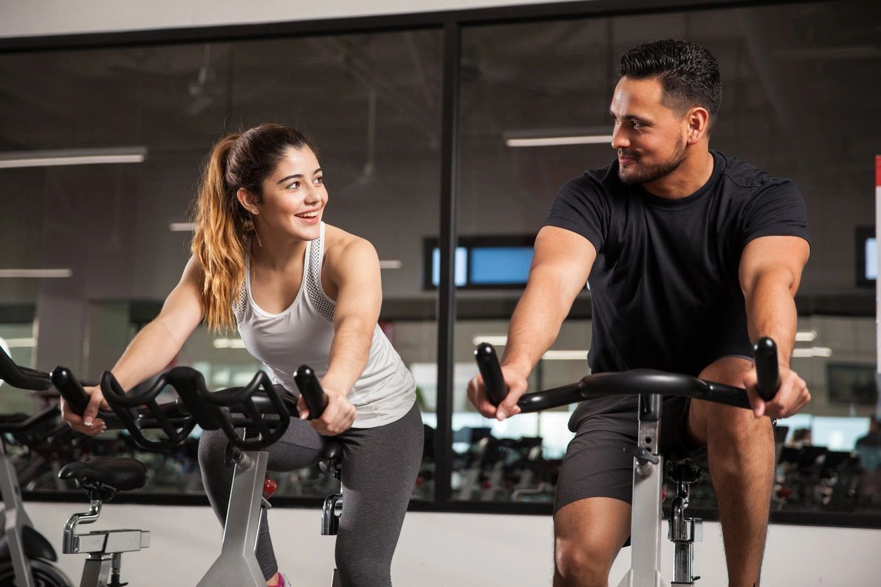 A man and a woman are riding exercise bikes in a gym.