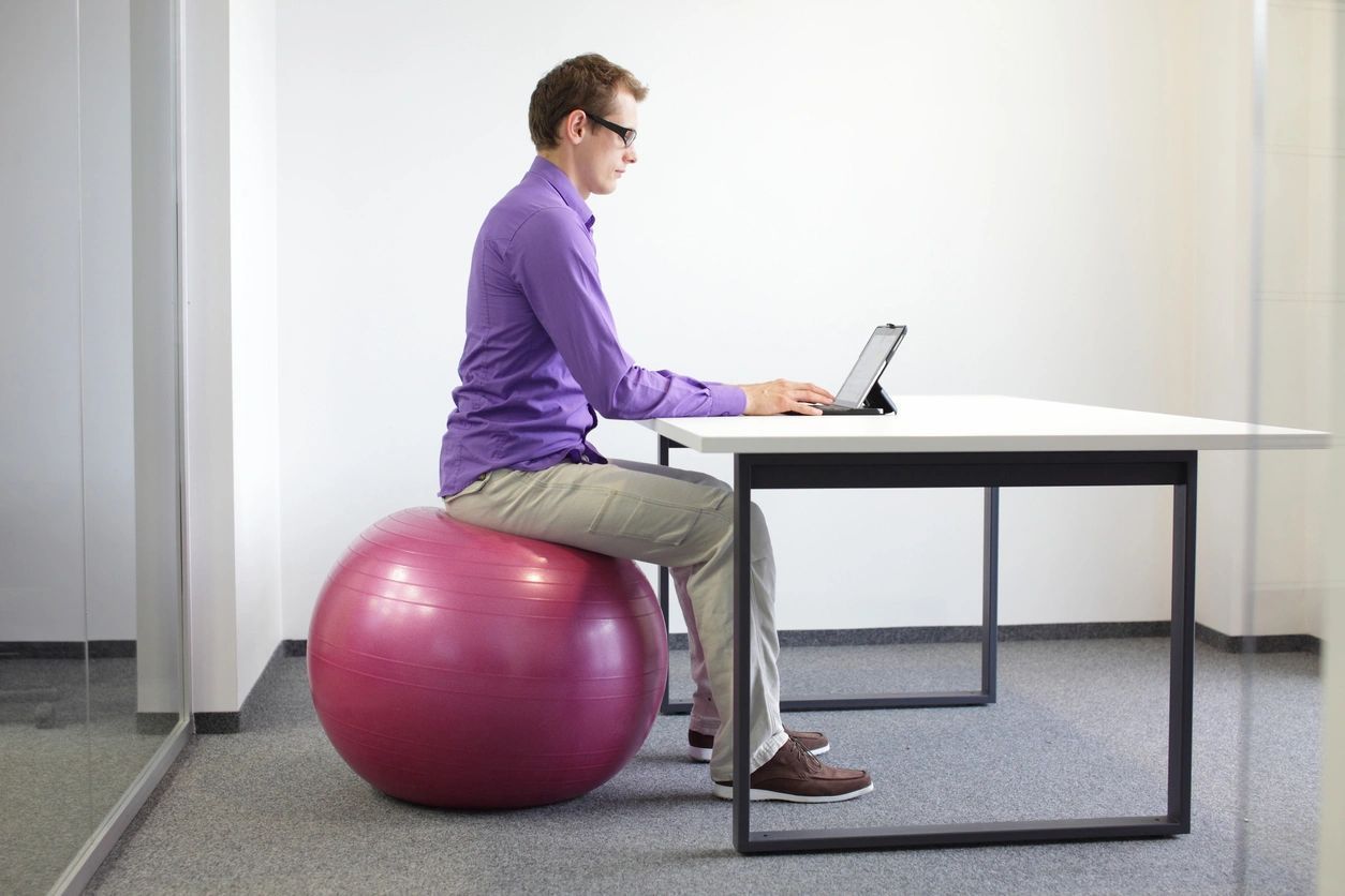 A man is sitting on an exercise ball while using a laptop.