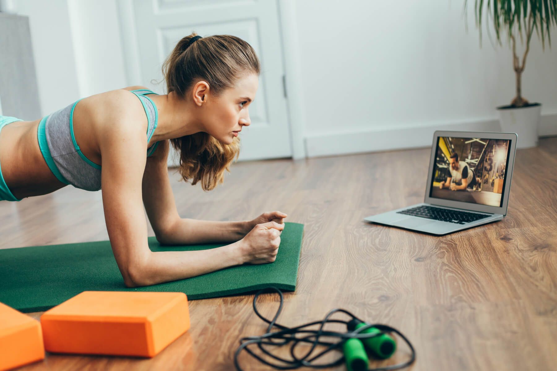 A woman is doing a plank on a yoga mat in front of a laptop computer.