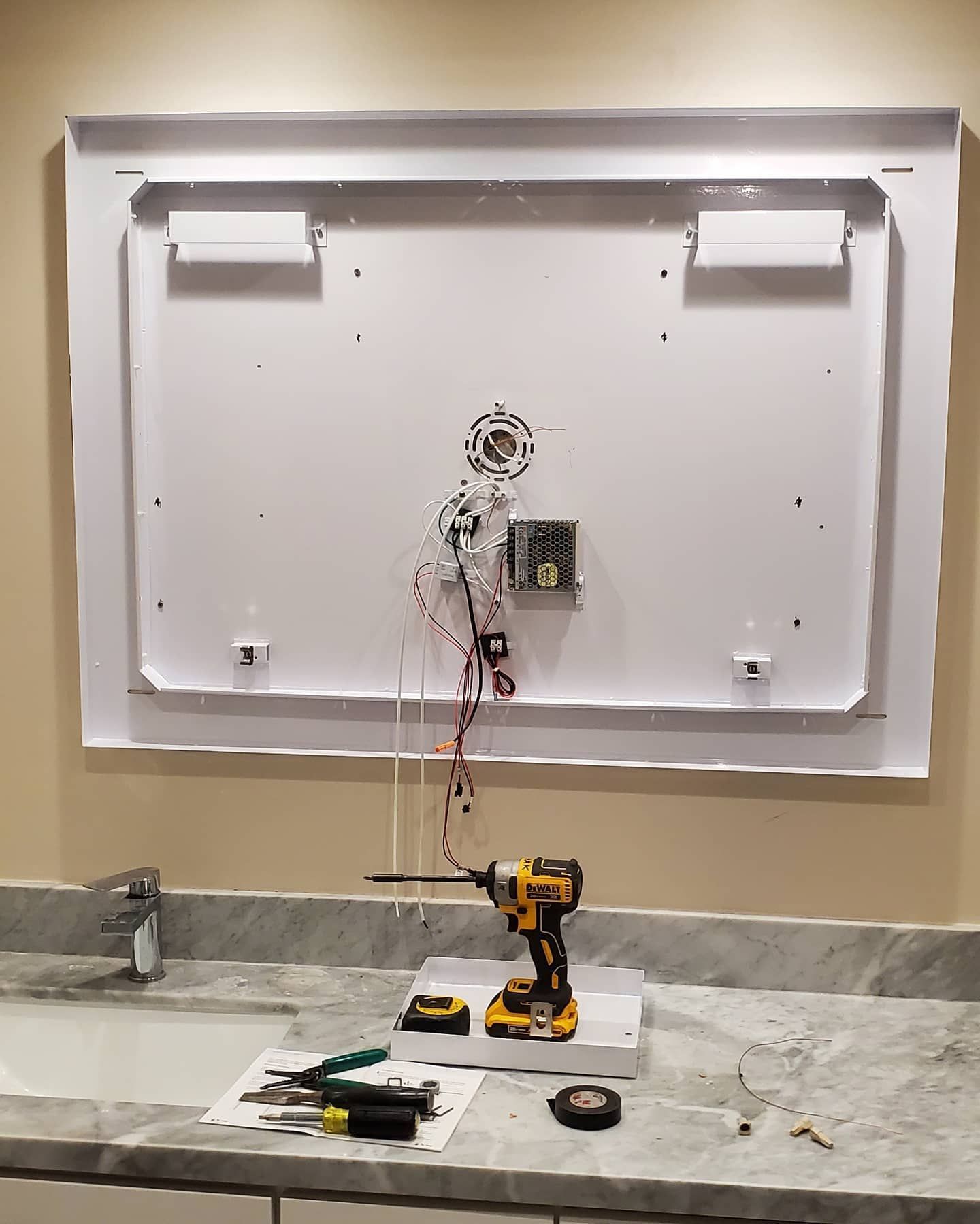 Back of a white framed vanity mirror with wiring exposed, over a bathroom countertop with tools.