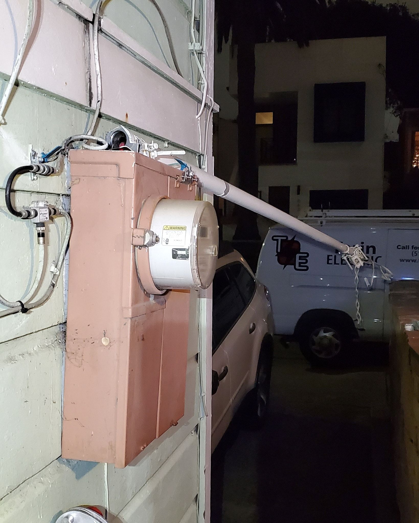 Electrical panel on a building's exterior with conduit extending to the right. A car and van are nearby.