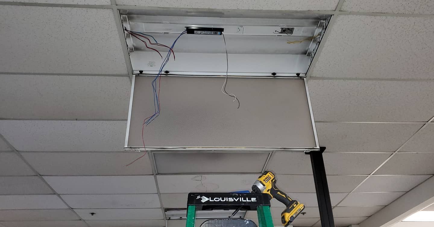 Fluorescent light fixture removed from ceiling, wires exposed. A ladder supports a drill below.