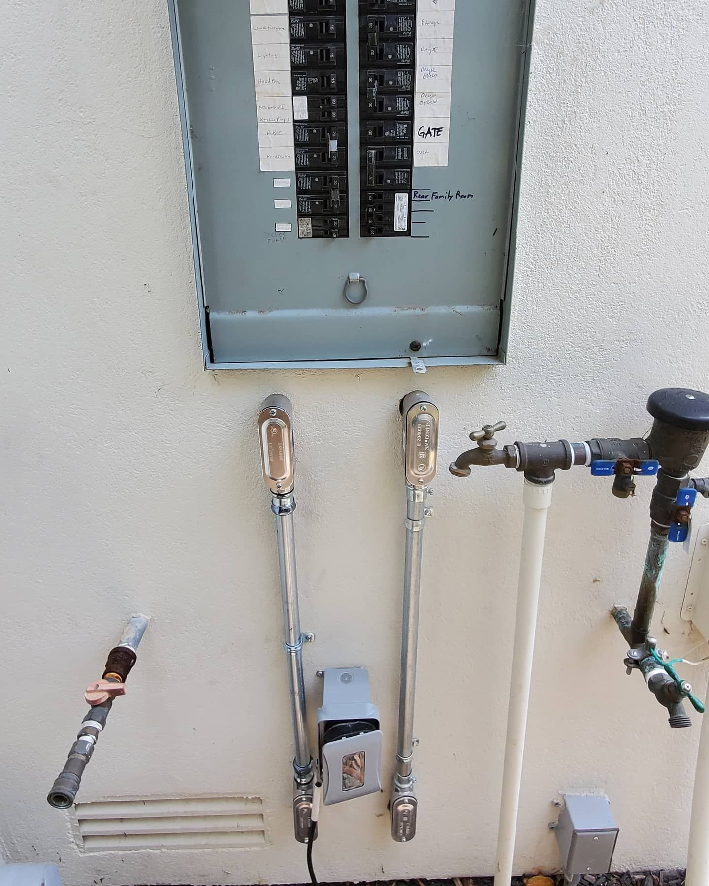Electrical panel, water pipes, and spigot mounted on a stucco wall.