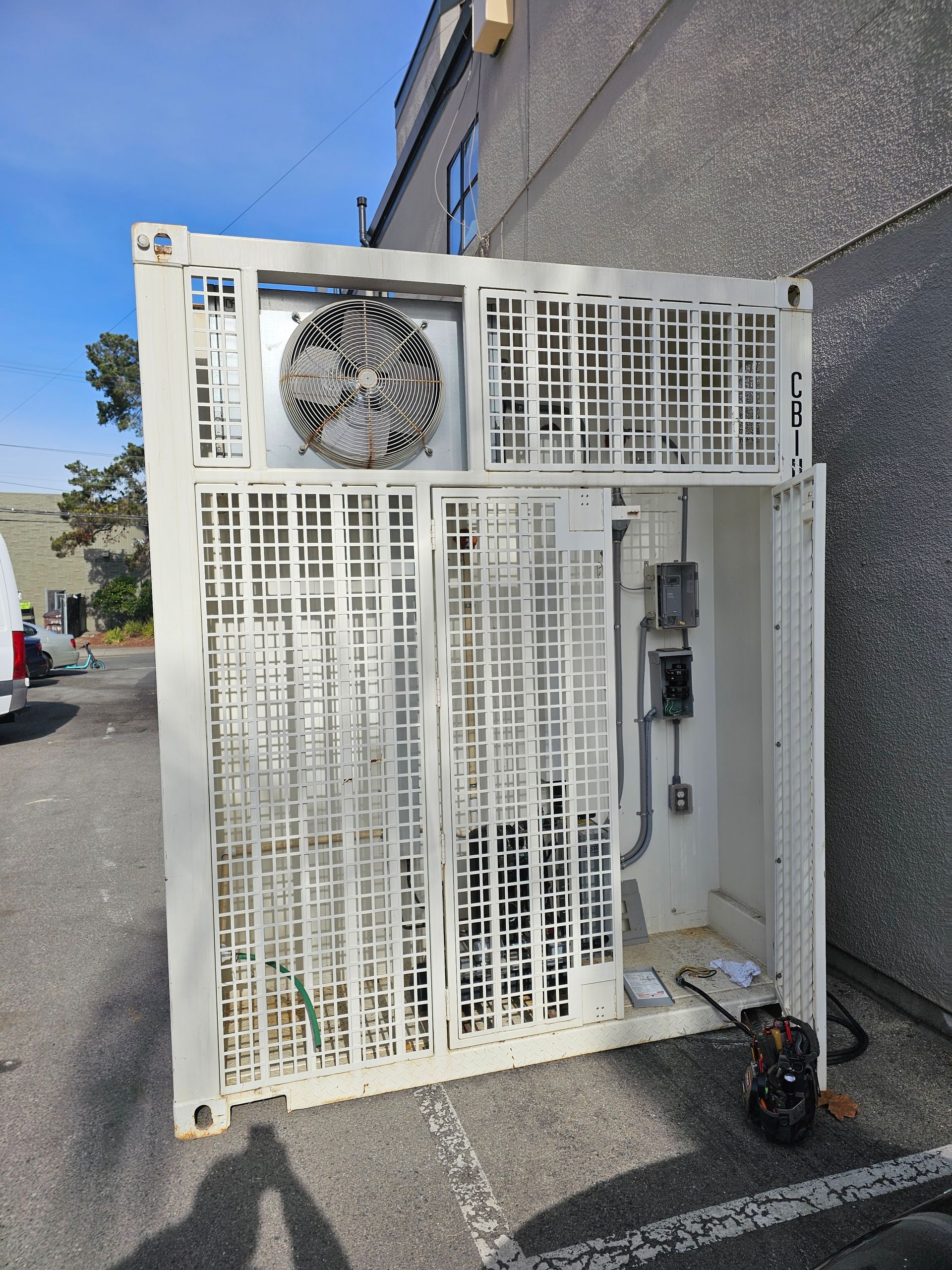 White metal industrial unit with open panels against a building. A fan and electrical components are visible.