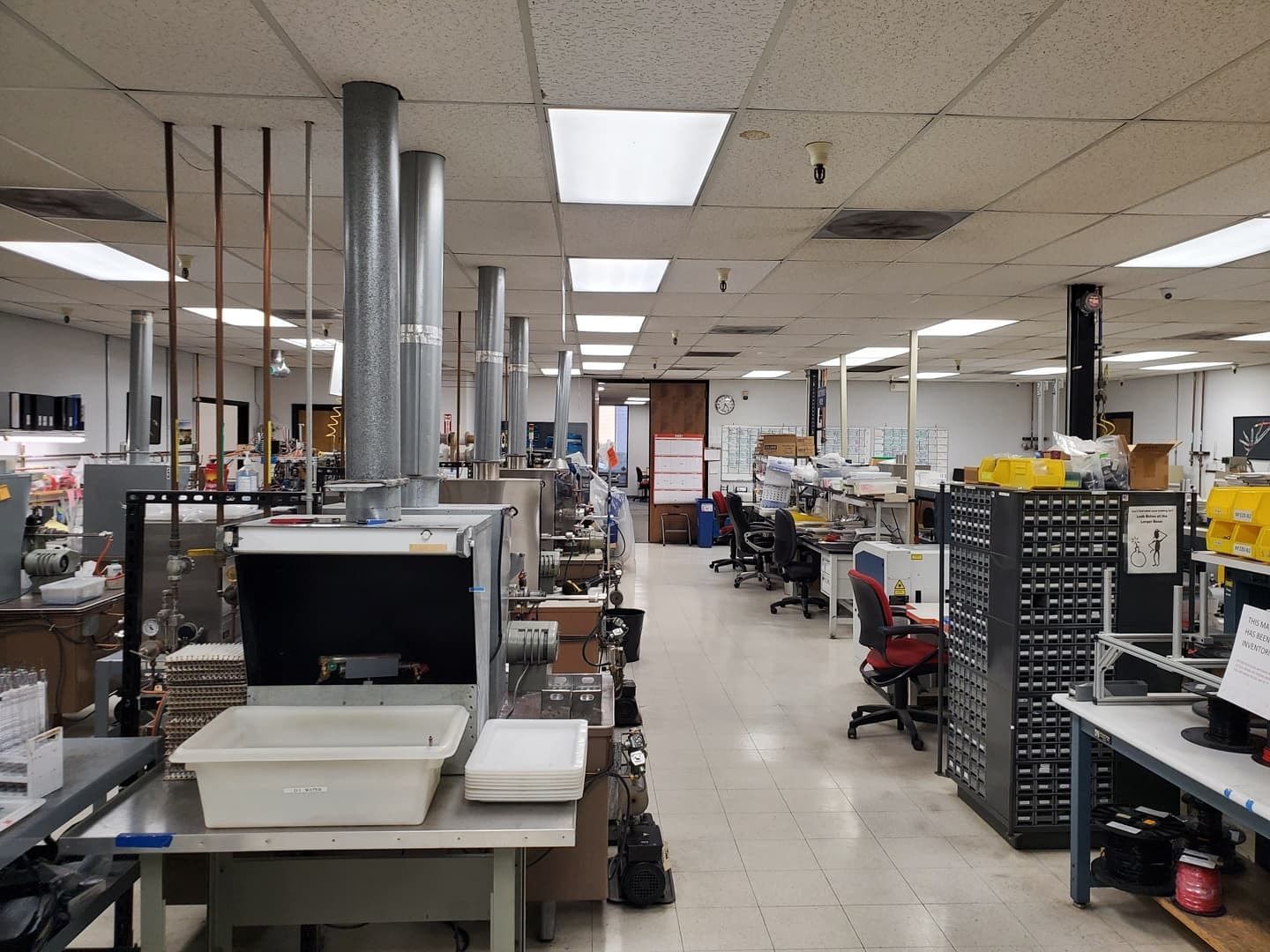 An industrial workshop with work stations, equipment, and ventilation systems. Fluorescent lighting illuminates the space.
