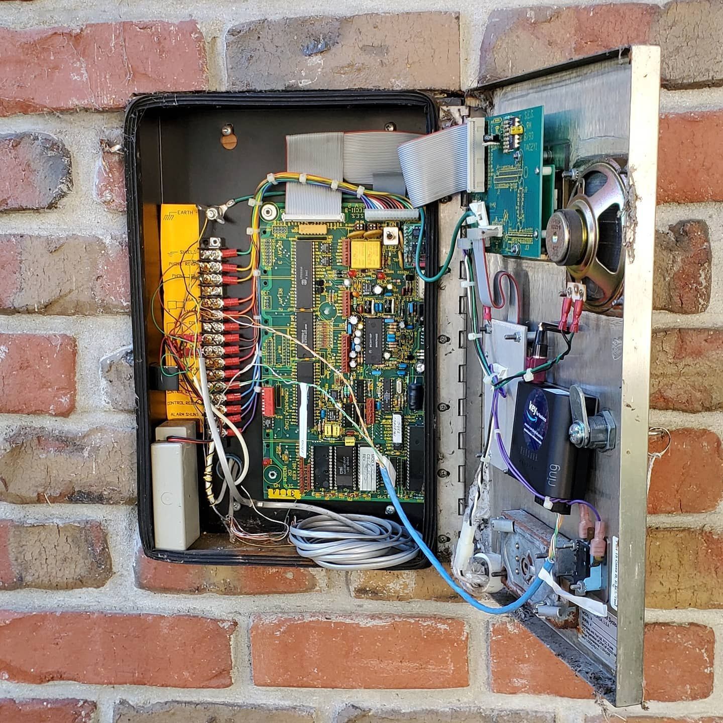 Open alarm system panel mounted on a brick wall, revealing circuit boards, wiring, and speaker.
