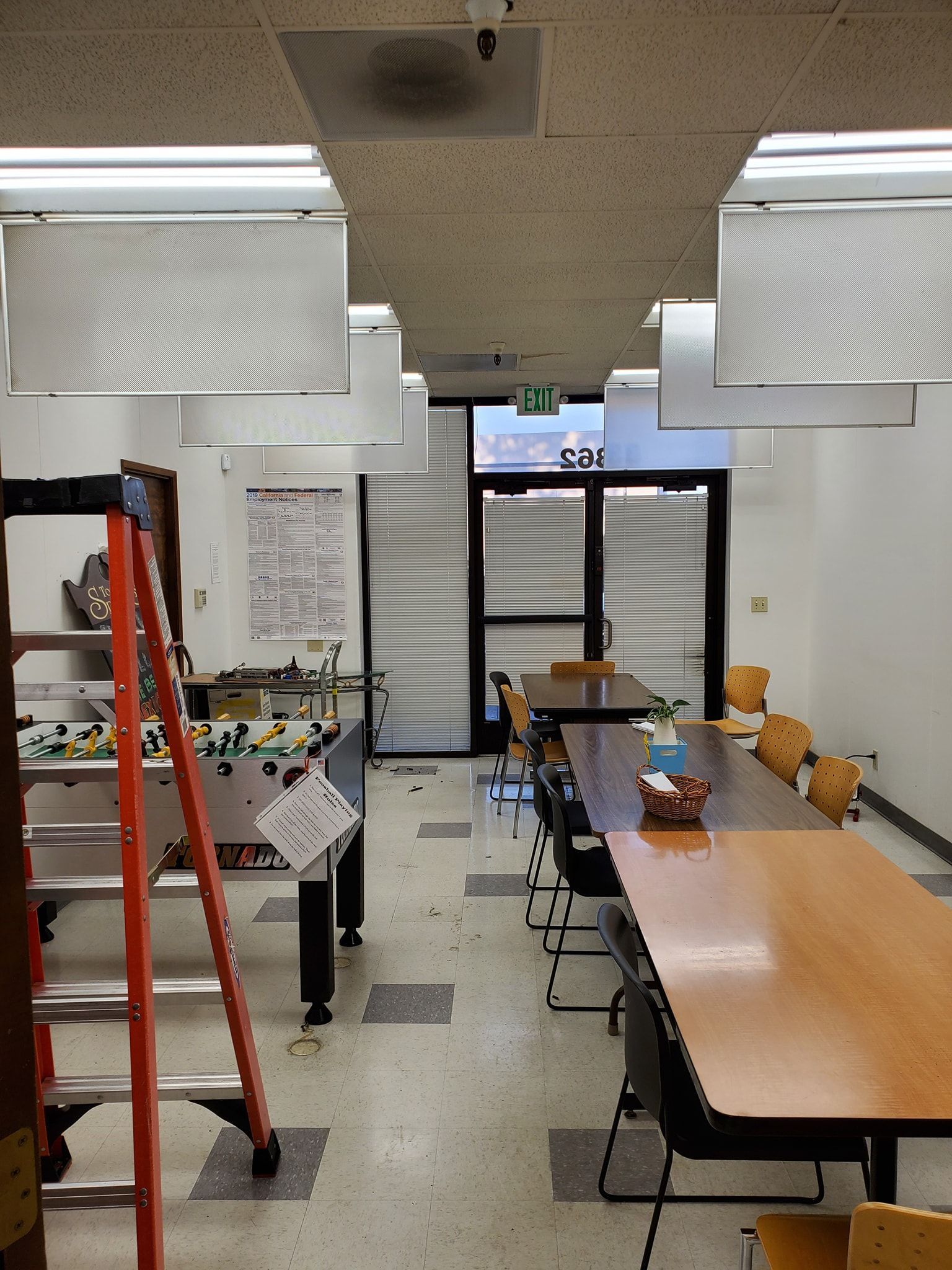 Interior of a room with tables and chairs, a foosball table, a ladder, and a door to the outside.
